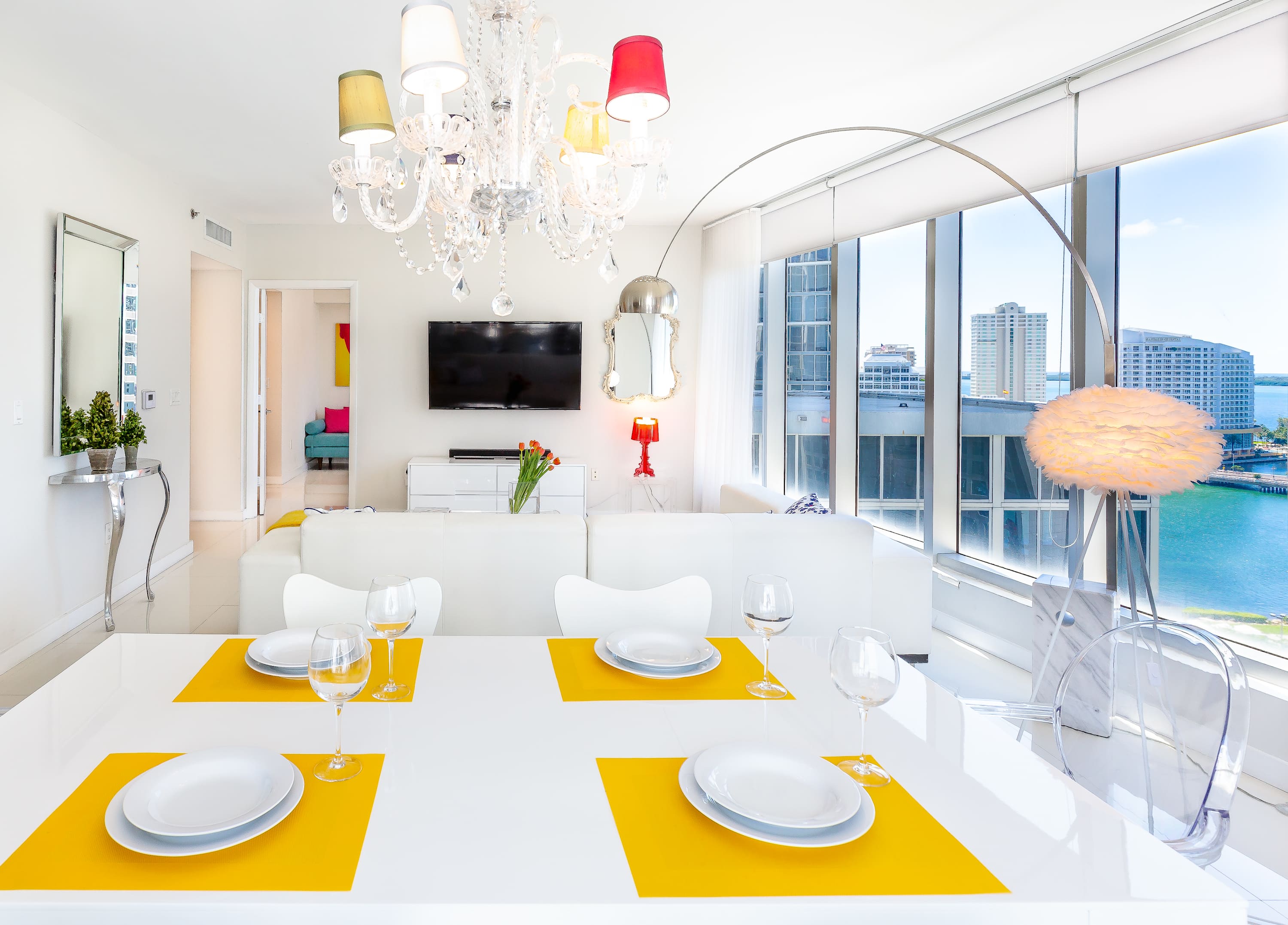 Modern dining area with a white table set with yellow placemats, clear chairs, and a vibrant chandelier. The space opens to a living area and offers stunning ocean and city views through large windows.
