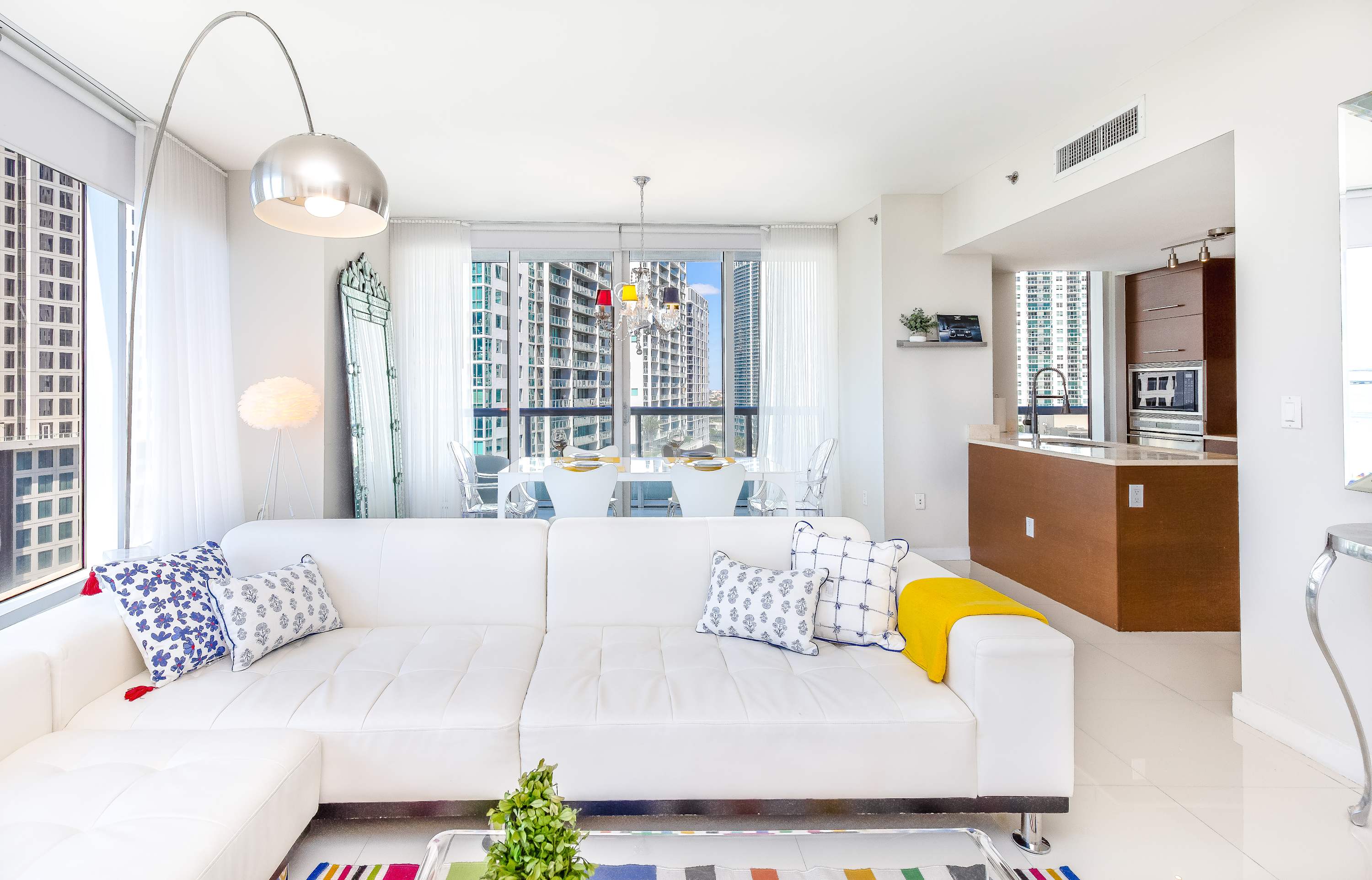 Spacious living room with a white sectional sofa, a clear coffee table, and a colorful striped rug, flowing into a dining area with a white table and clear chairs. The open-concept space features large windows with city views and a partial view of the modern kitchen island.
