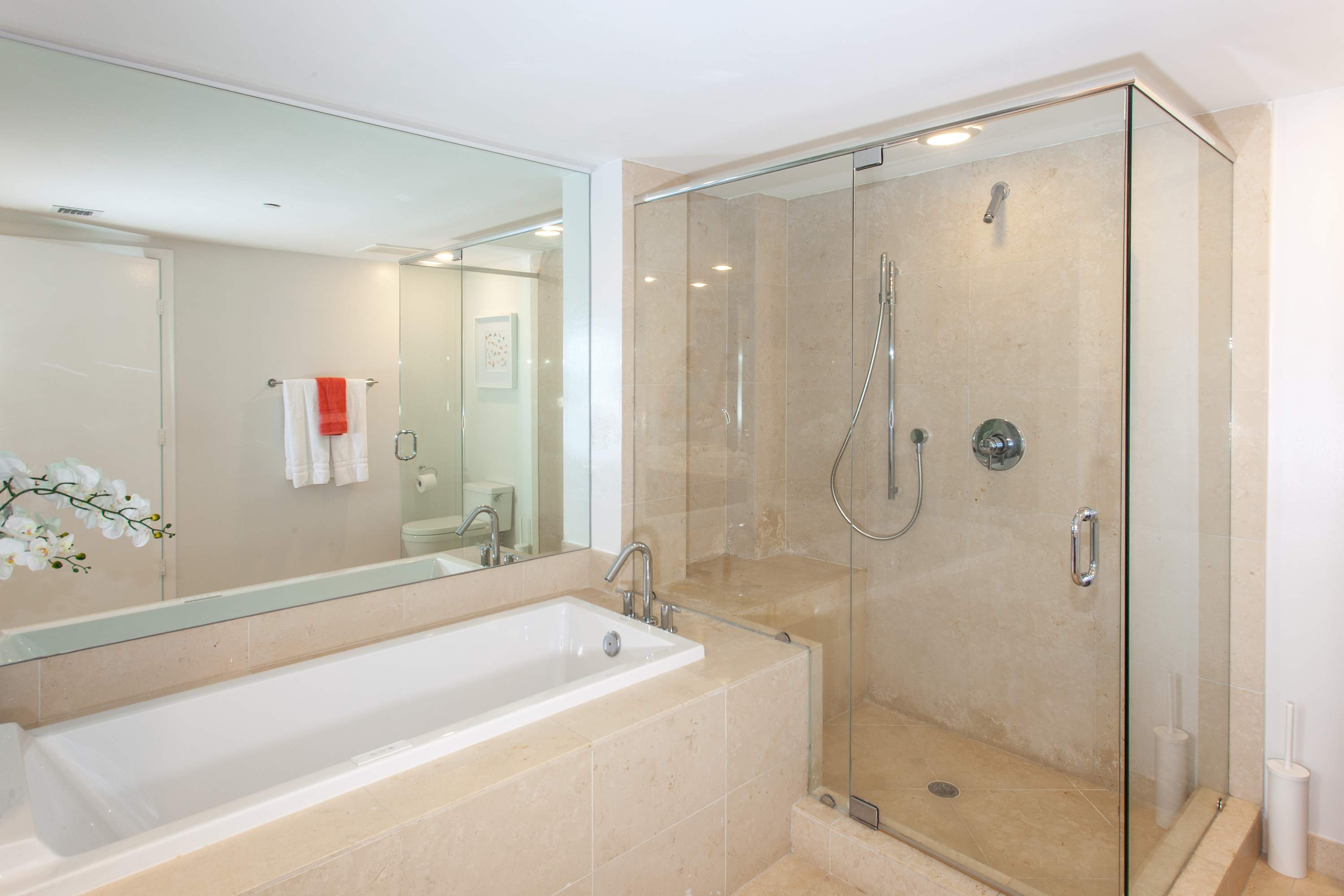 Modern bathroom featuring a large built-in bathtub and a glass-enclosed walk-in shower, both surrounded by light-colored stone tiles. A large mirror reflects the opposite wall.