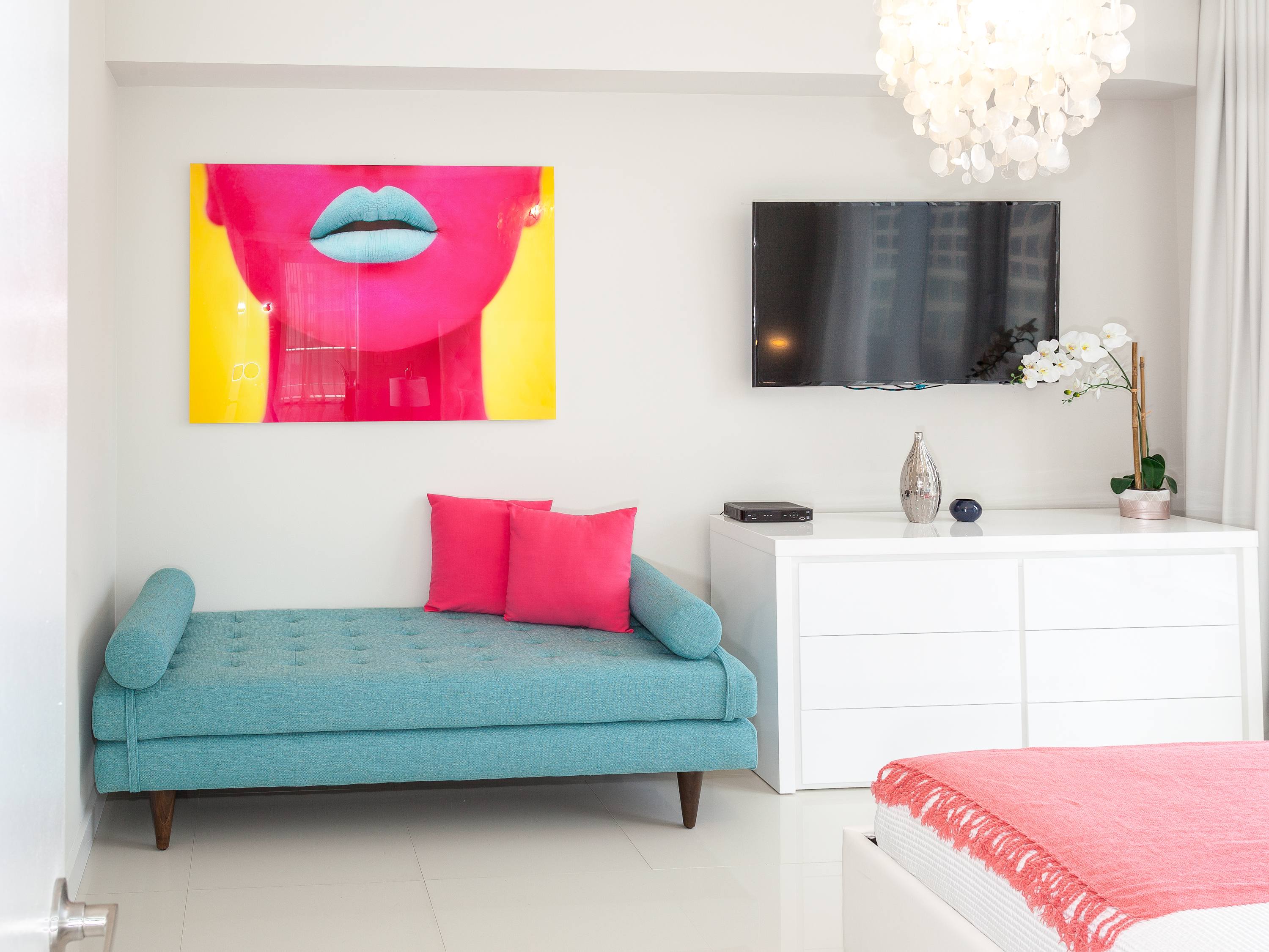 Vibrant bedroom featuring a bright pink and yellow pop art piece above a turquoise daybed with hot pink pillows. A wall-mounted TV is positioned above a white dresser, and a shell-like chandelier hangs from the ceiling, adding a unique touch.