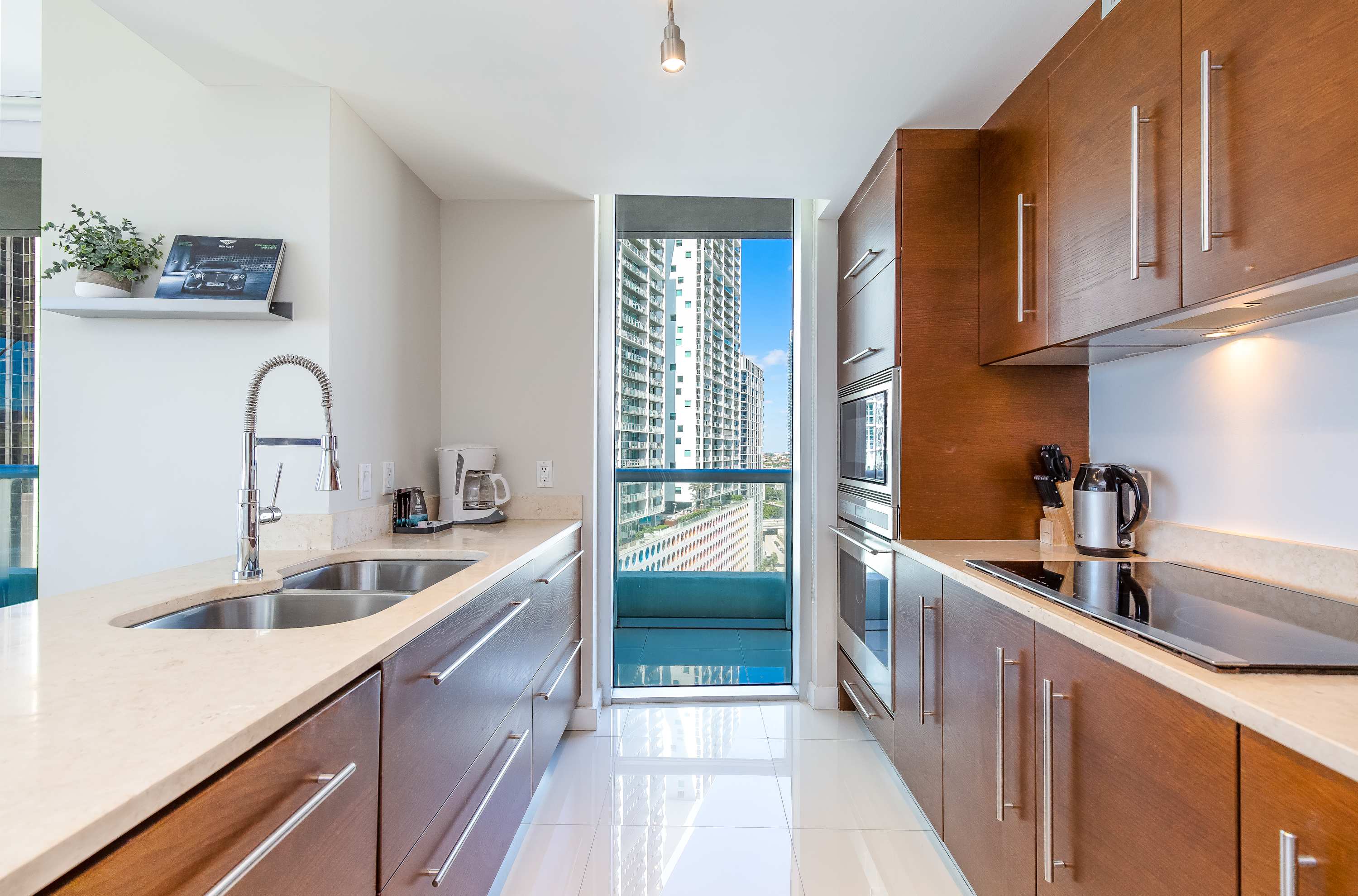 Modern galley kitchen with sleek wooden cabinetry, light beige countertops, and stainless steel appliances including a double sink and an induction cooktop. A narrow window at the end offers a glimpse of city buildings.