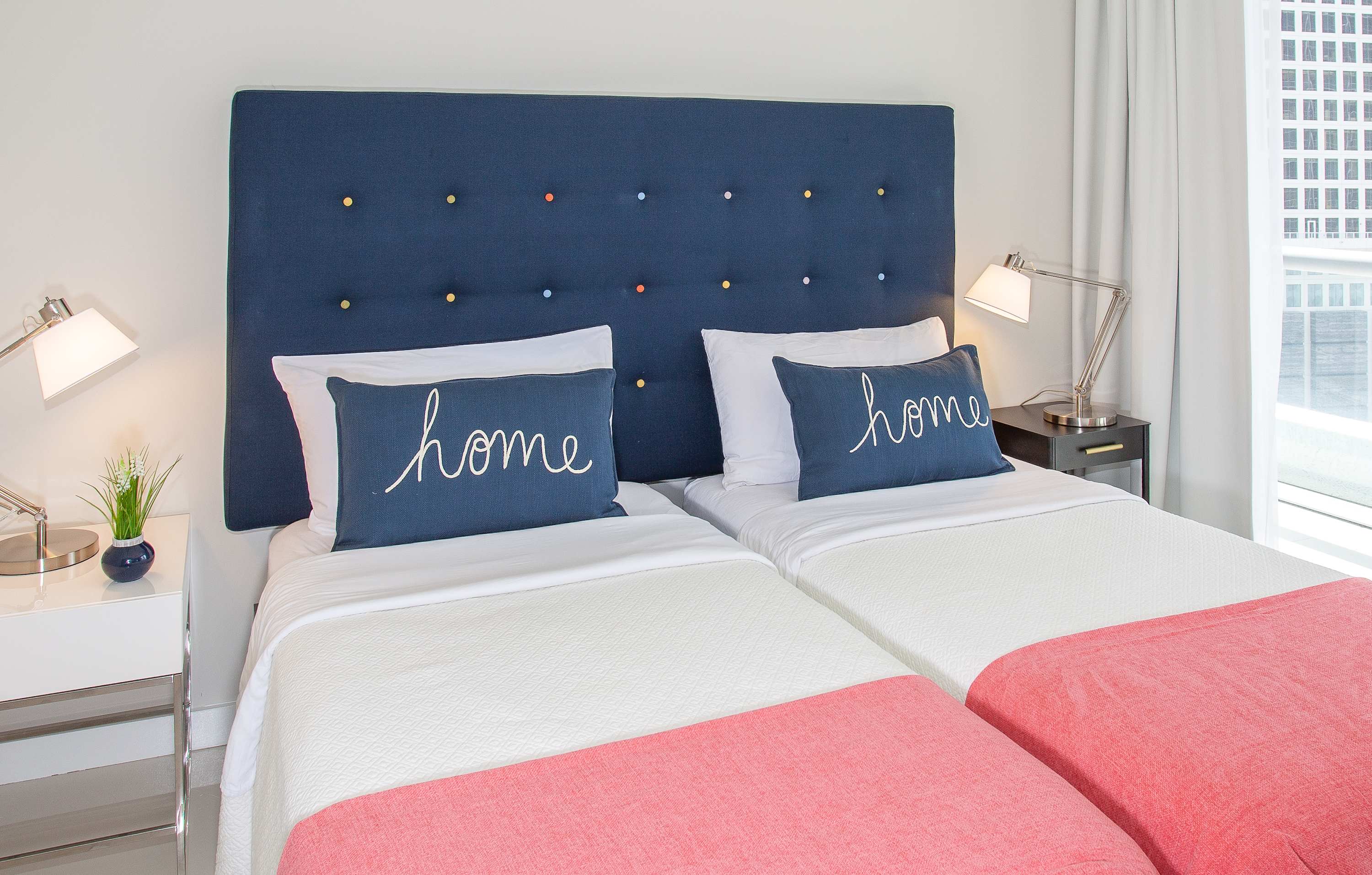 Cozy bedroom featuring two twin beds with navy blue headboards and 'home' embroidered pillows, flanked by modern nightstands with task lamps. White bedding with coral throws adds a pop of color to the bright space.