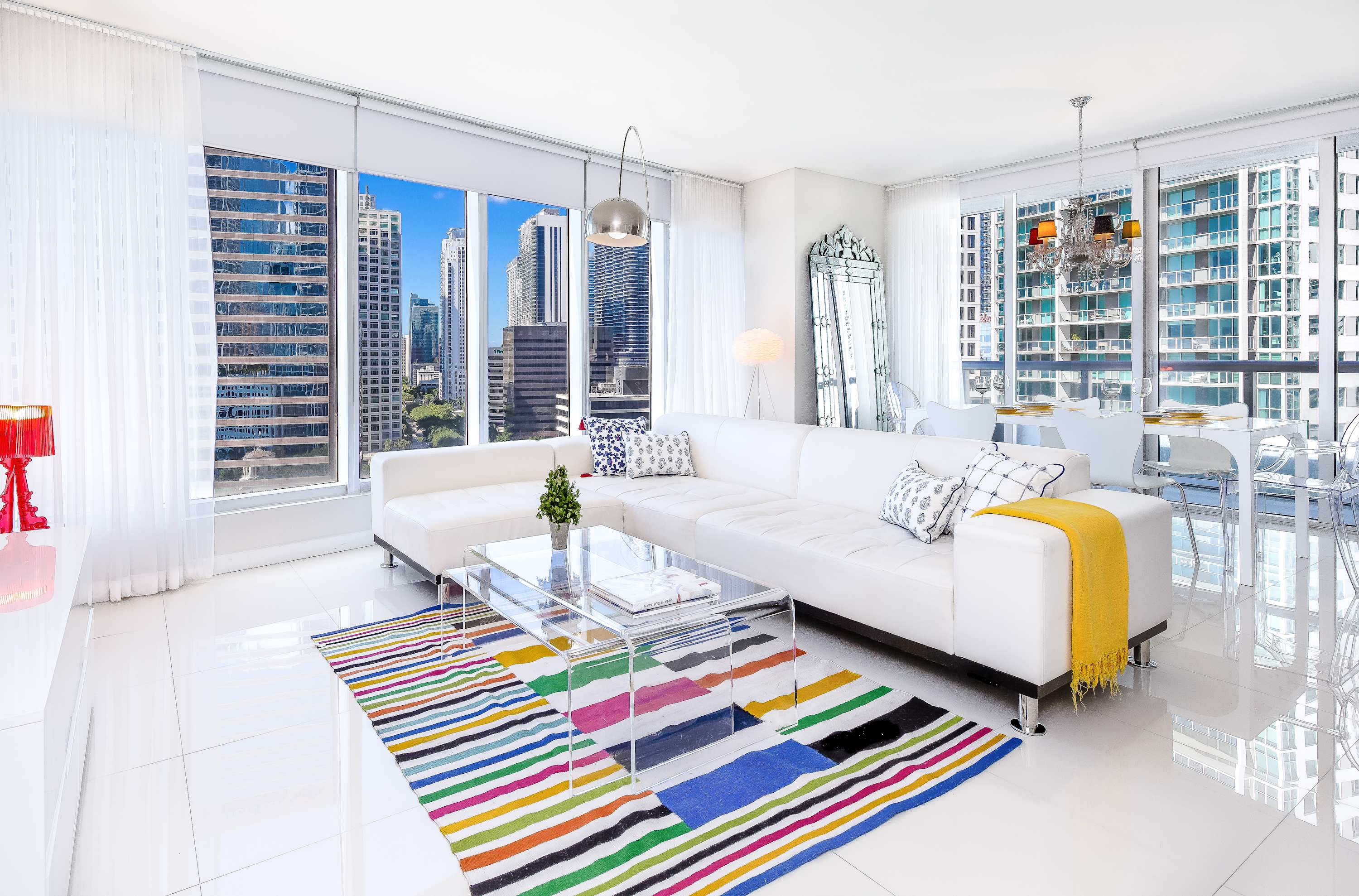 Bright and modern living room featuring a large white sectional sofa, a clear acrylic coffee table, and a vibrant striped rug. Floor-to-ceiling windows offer expansive city views, and the room is decorated with contemporary art and stylish lighting.