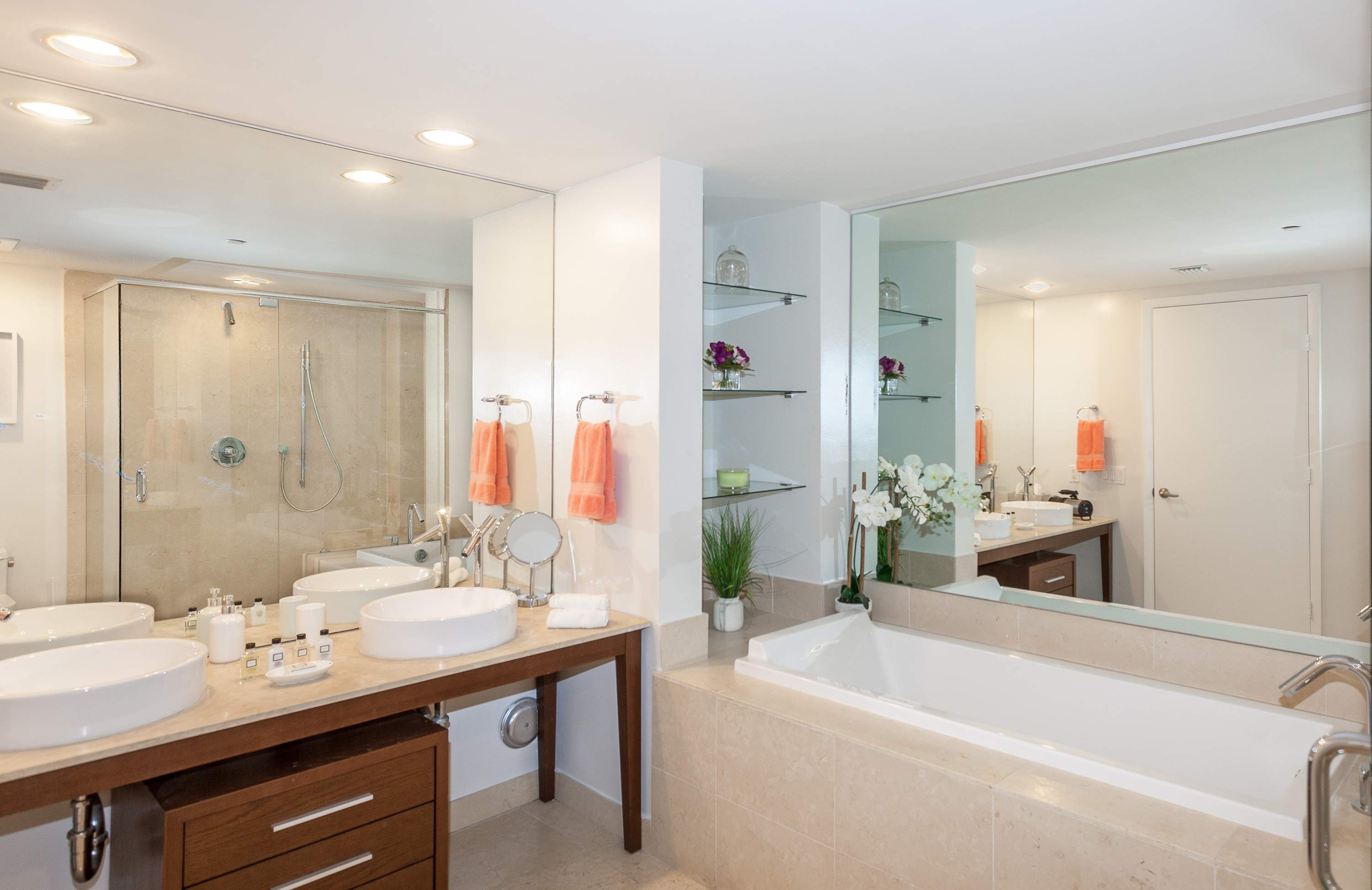 Spacious modern bathroom with a double vanity featuring vessel sinks on a light stone countertop, a large mirror, and a built-in bathtub. Shelving with decorative items is visible in the background.
