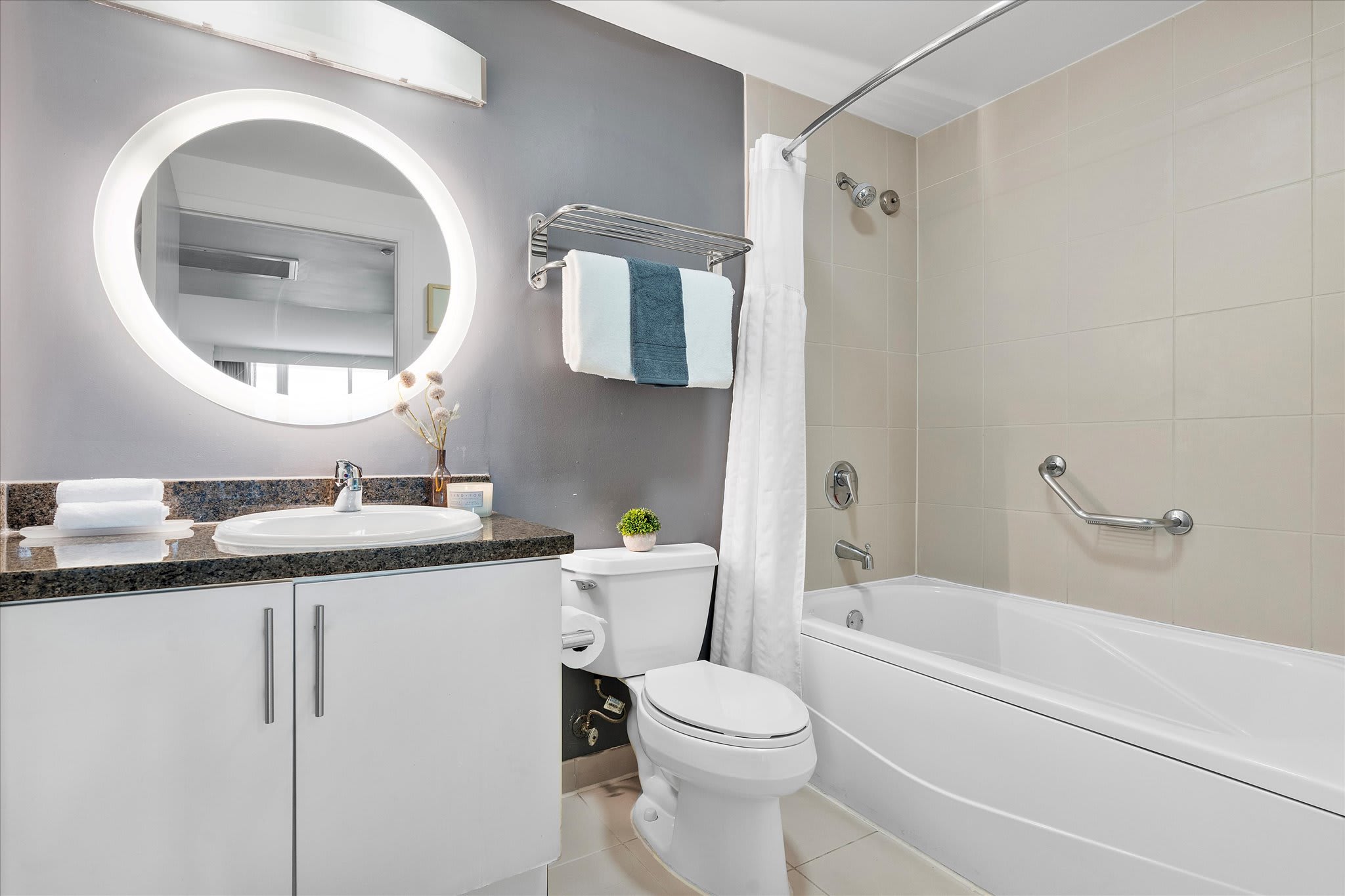 A clean, modern bathroom with a white vanity, circular illuminated mirror, toilet, and a bathtub with a shower. The walls are light beige and grey, with a towel rack.