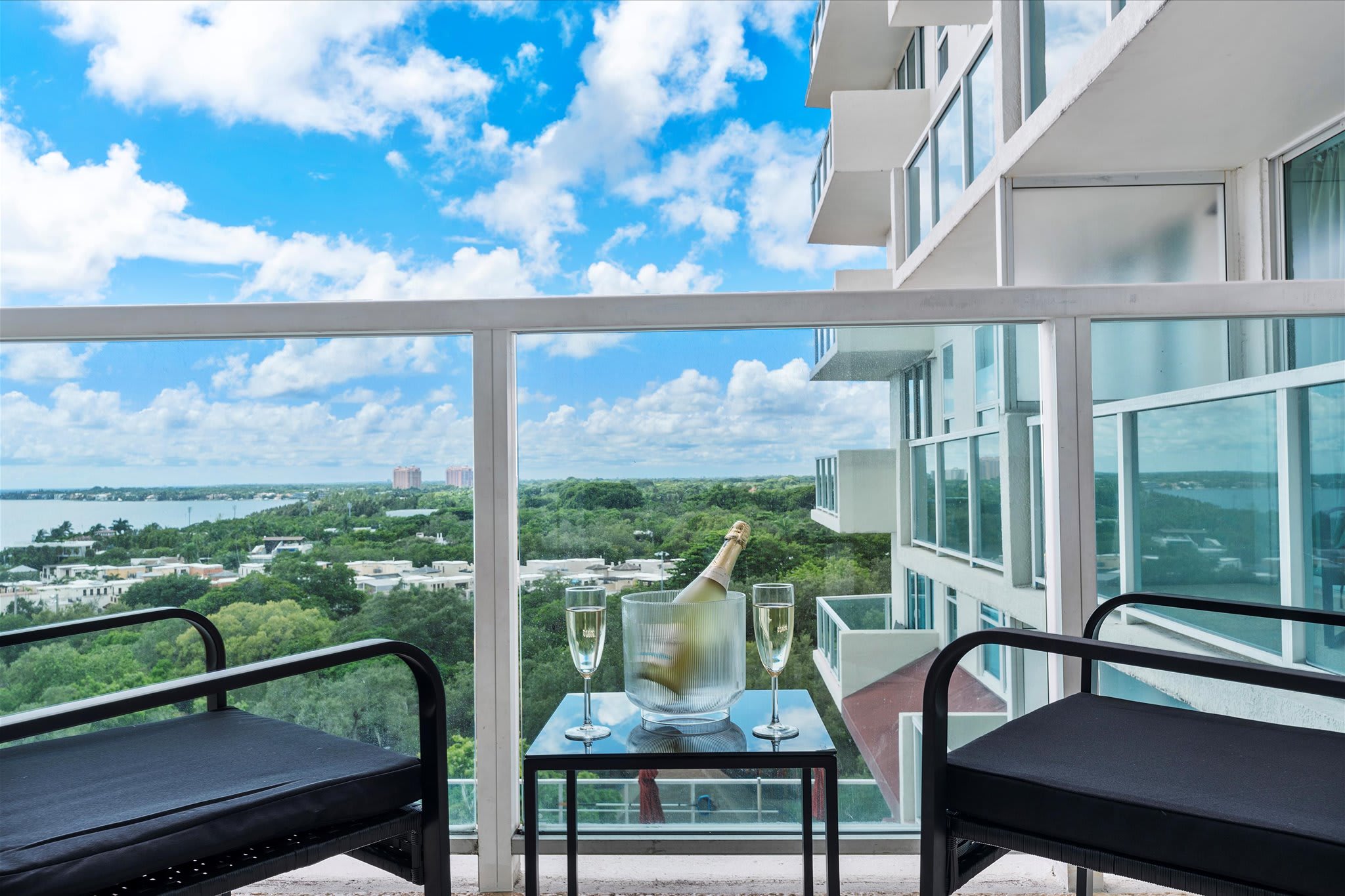 High-rise balcony with glass railings, two black chairs, and a small table holding a bucket with a champagne bottle and two flutes. The view overlooks lush greenery and a bay under a blue sky.