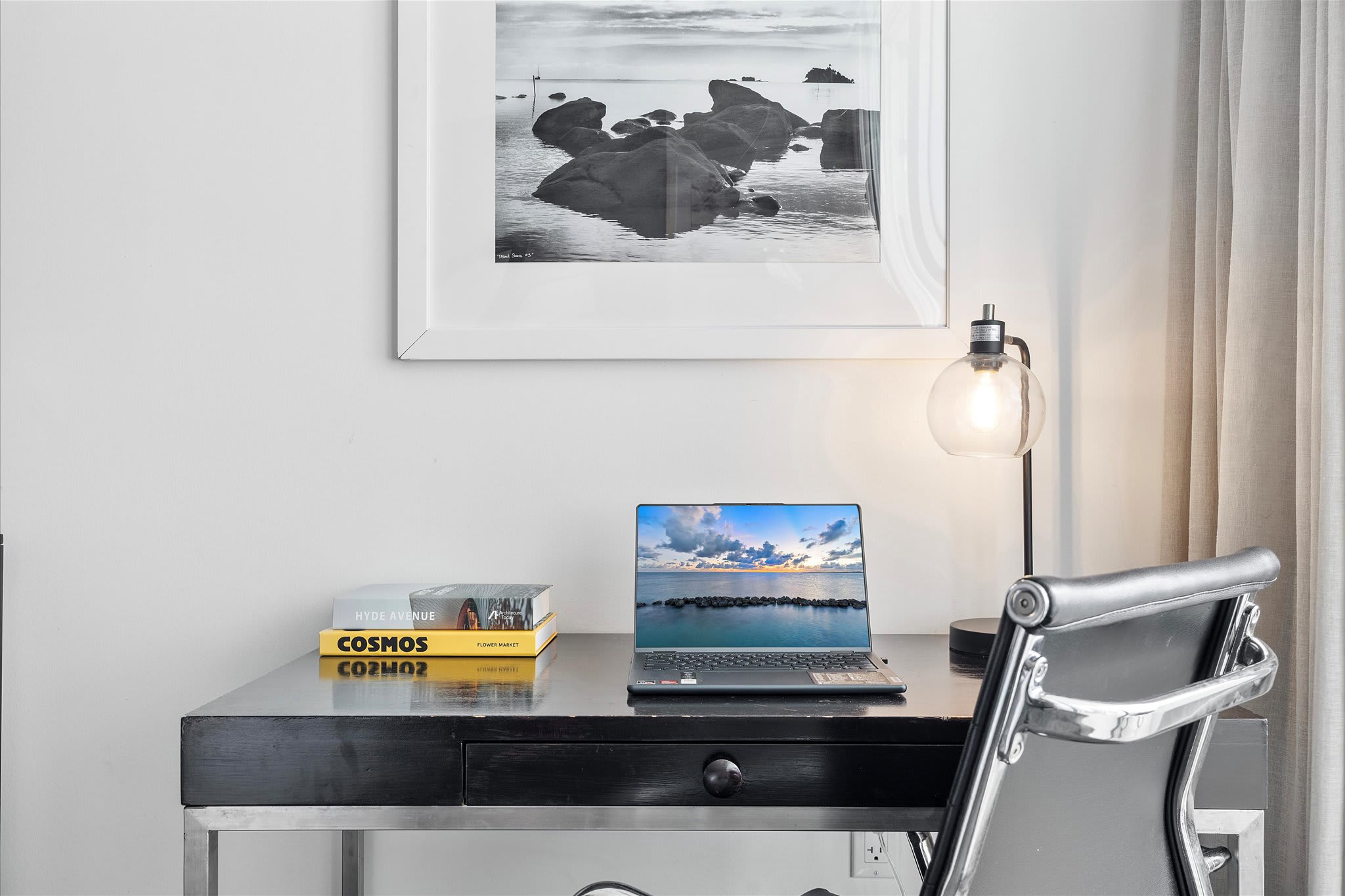 A dedicated workspace within the studio, featuring a black desk, a laptop, and a modern desk lamp. A framed black and white photo hangs above, with books stacked on the desk.