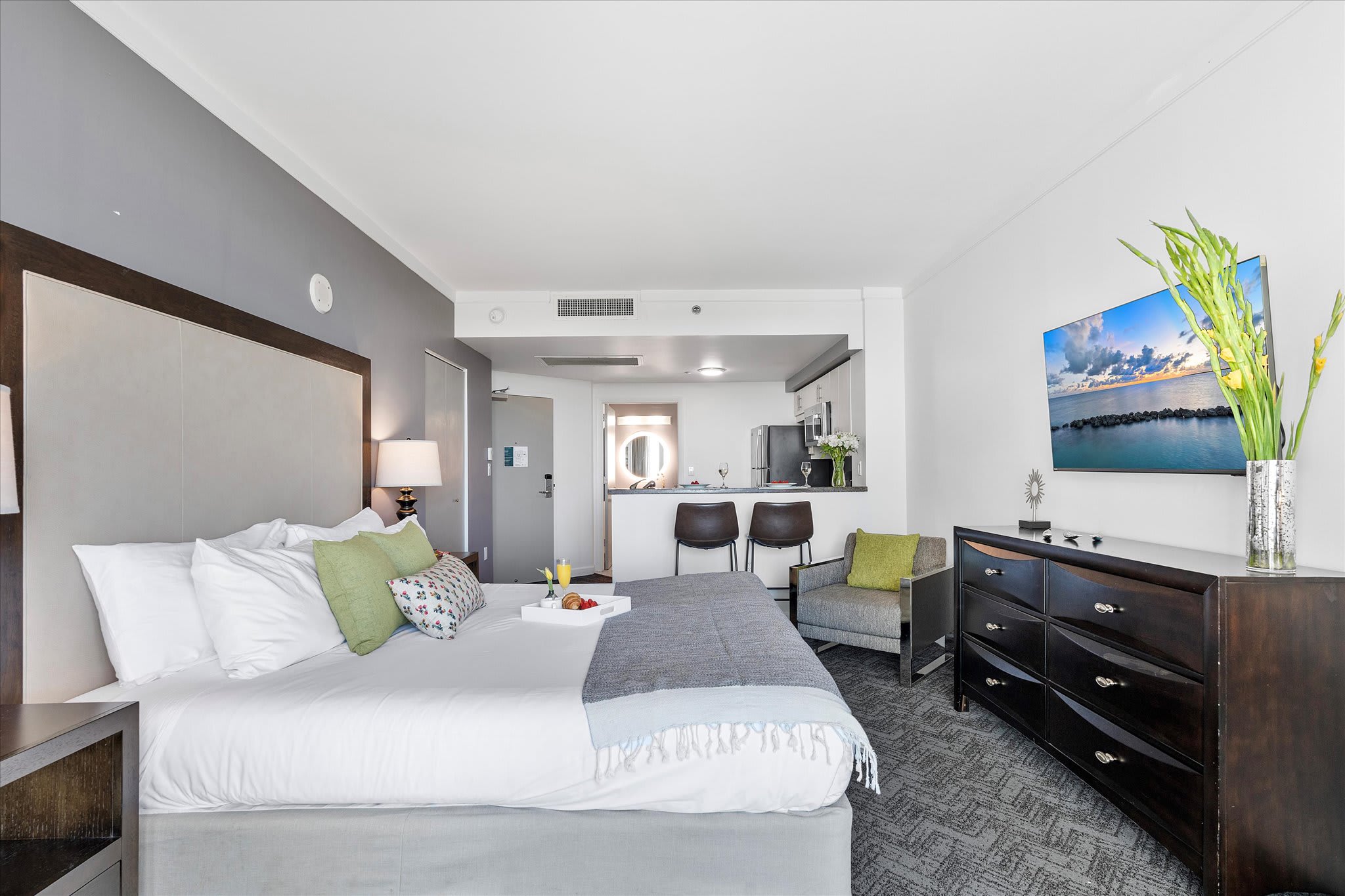 A studio apartment layout showing the king-sized bed with a large headboard, a sitting chair, and the open-concept kitchen area with a breakfast bar in the background.