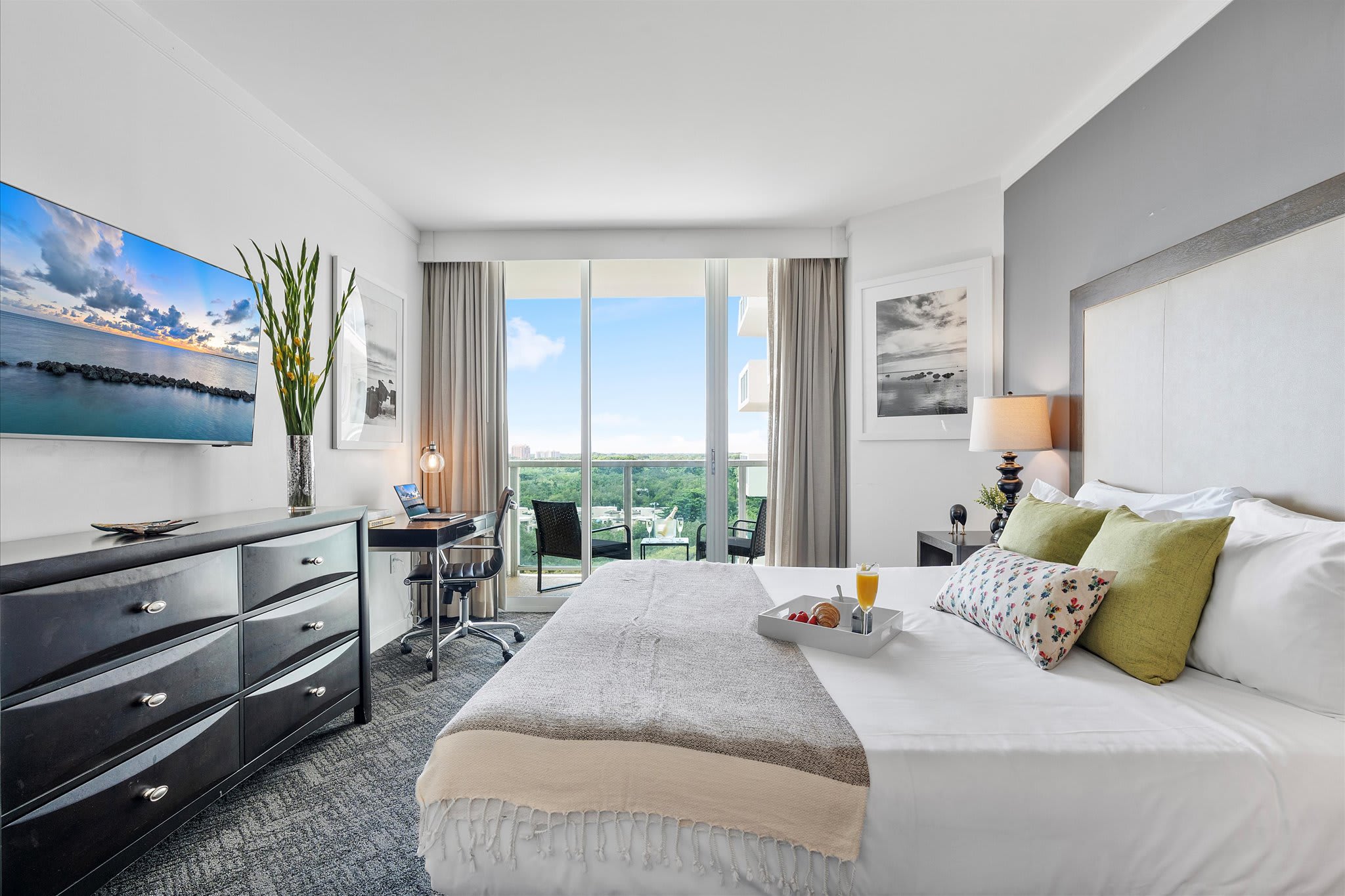 Modern studio bedroom featuring a king bed, a dark wood dresser with a TV, and a desk area. Floor-to-ceiling windows lead to a balcony with a view of the bay and greenery.