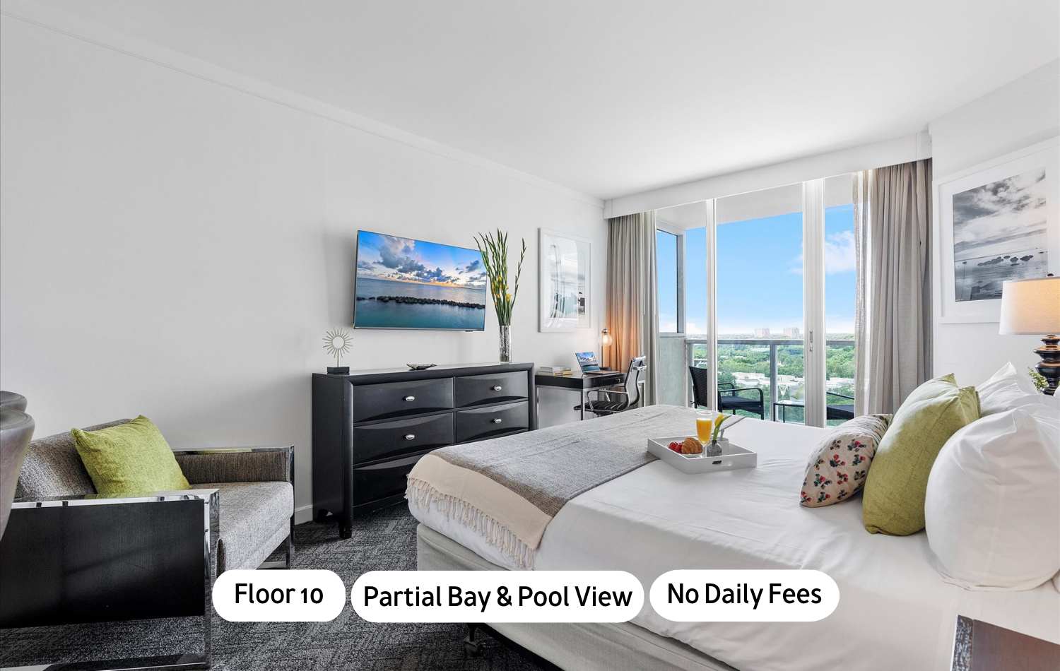 Modern studio bedroom with a king bed, dark wood dresser with a flat-screen TV, and a small desk area. Floor-to-ceiling windows open to a balcony with partial bay and pool views.
