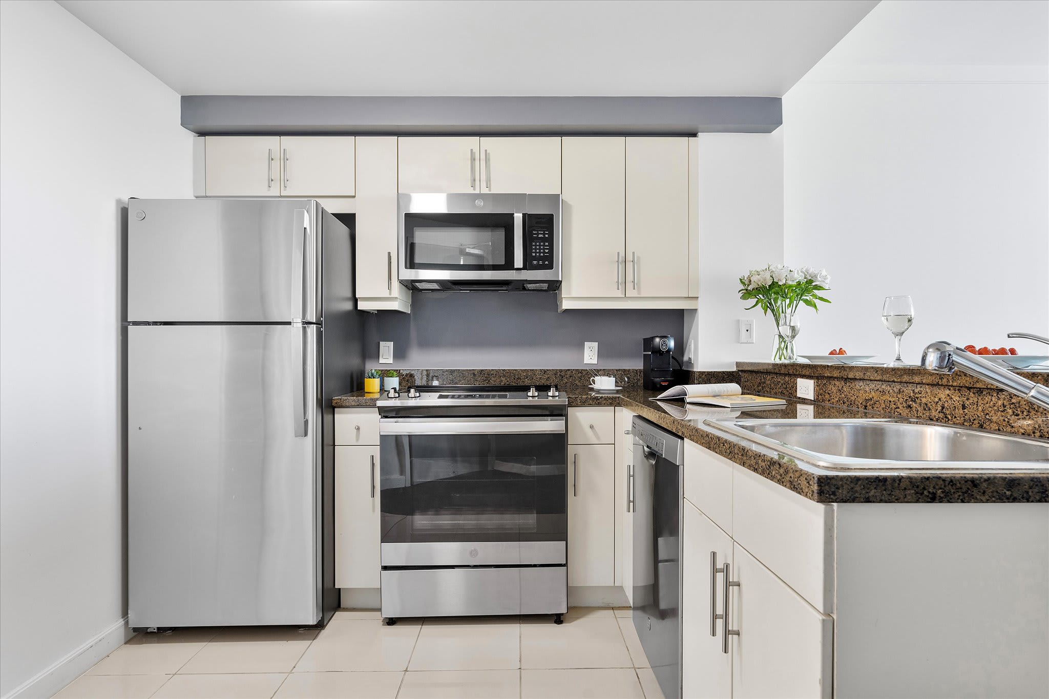 Compact modern kitchen with light cream cabinets, dark granite countertops, and stainless steel appliances including a refrigerator, oven, microwave, and dishwasher. A grey accent wall adds contrast.
