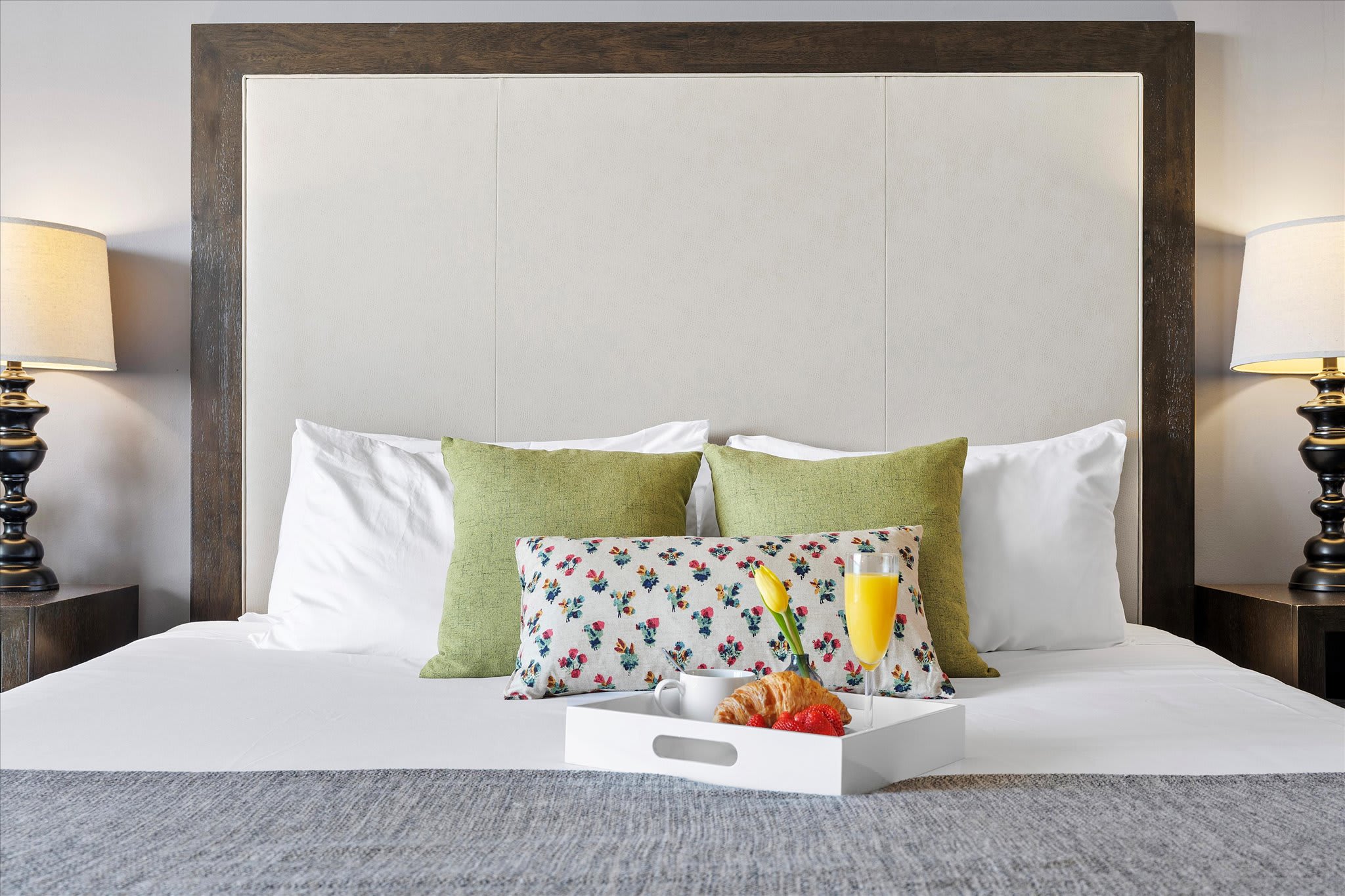 Close-up of a king-sized bed with a tall upholstered headboard, white and green pillows, and a decorative floral pillow. A white tray with breakfast items is placed on the bed.