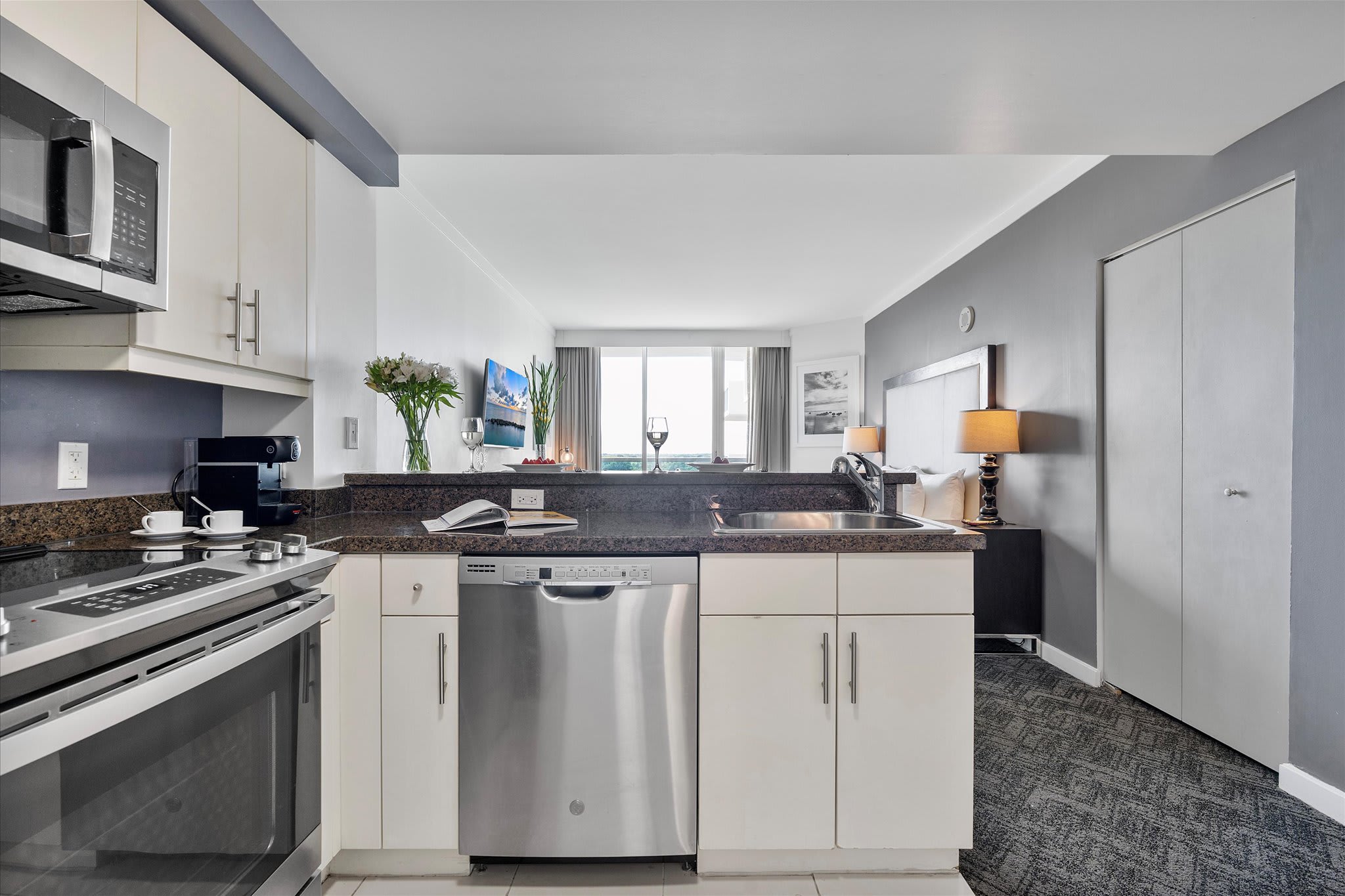 A modern kitchen with stainless steel appliances and granite countertops, featuring a breakfast bar with two stools. The view extends into the studio bedroom area.