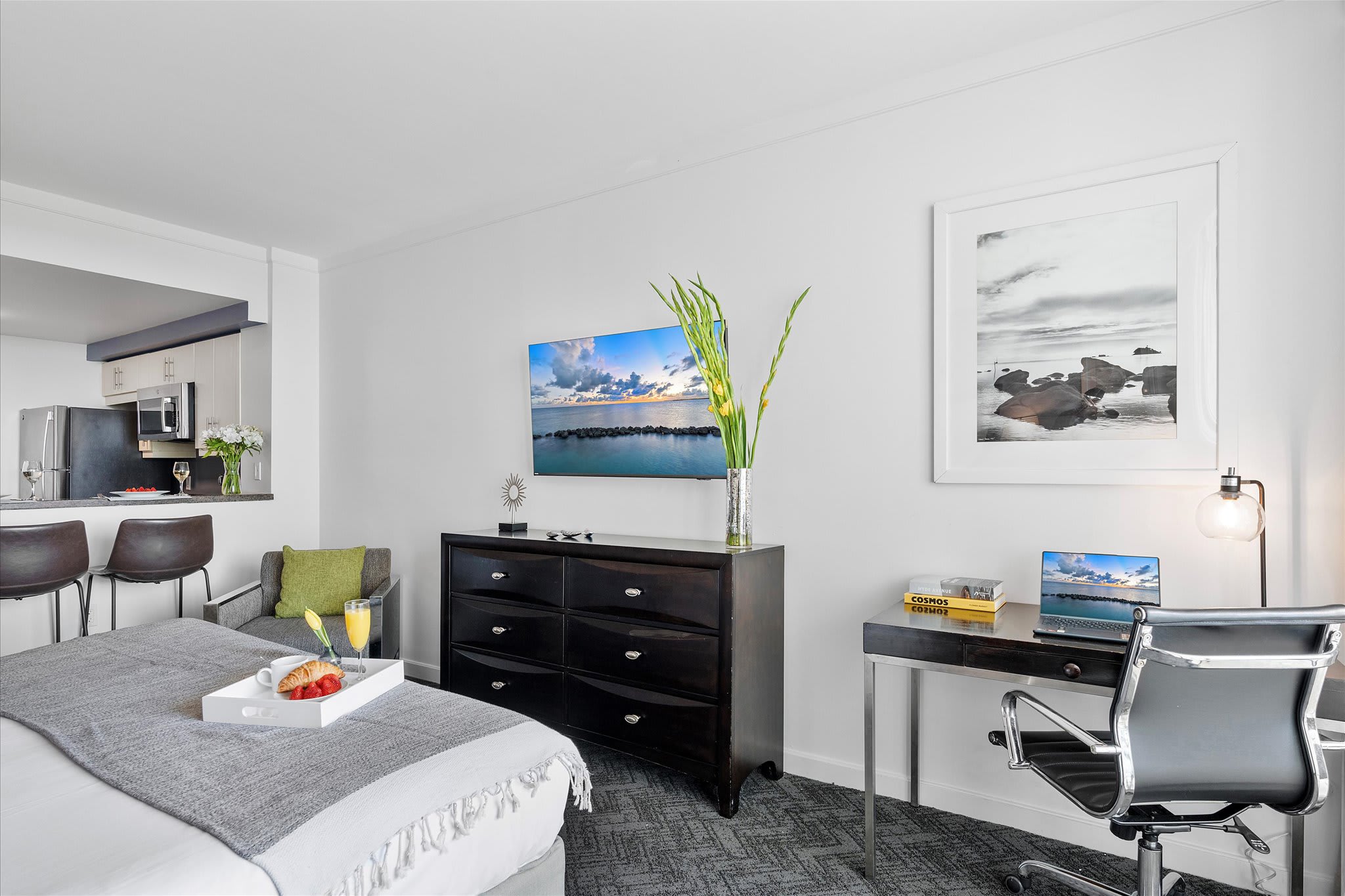 View of the studio bedroom from the foot of the bed, showing the dresser with a TV, a desk, and a partial view of the kitchen area with bar seating in the background.