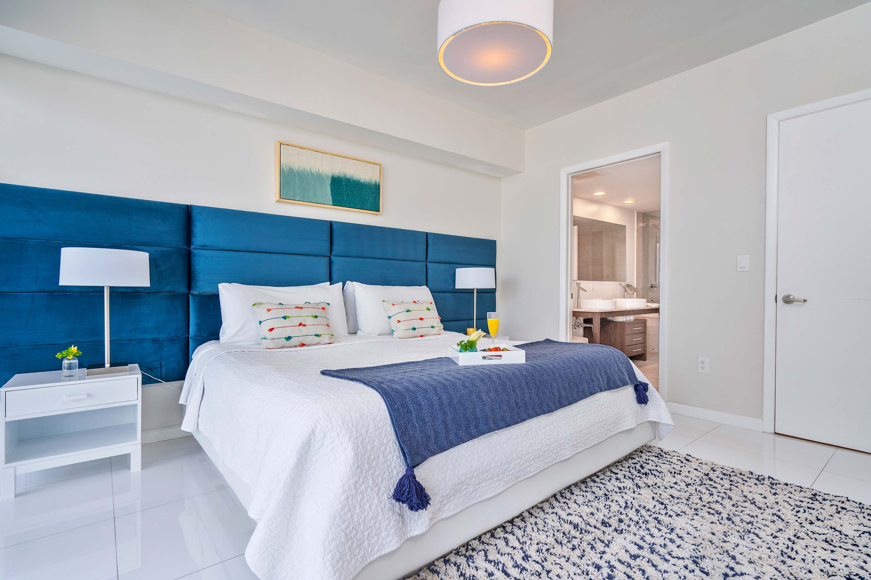 Elegant bedroom featuring a large white bed with a plush blue upholstered headboard, white nightstands, and an open doorway revealing a glimpse of the en-suite bathroom.