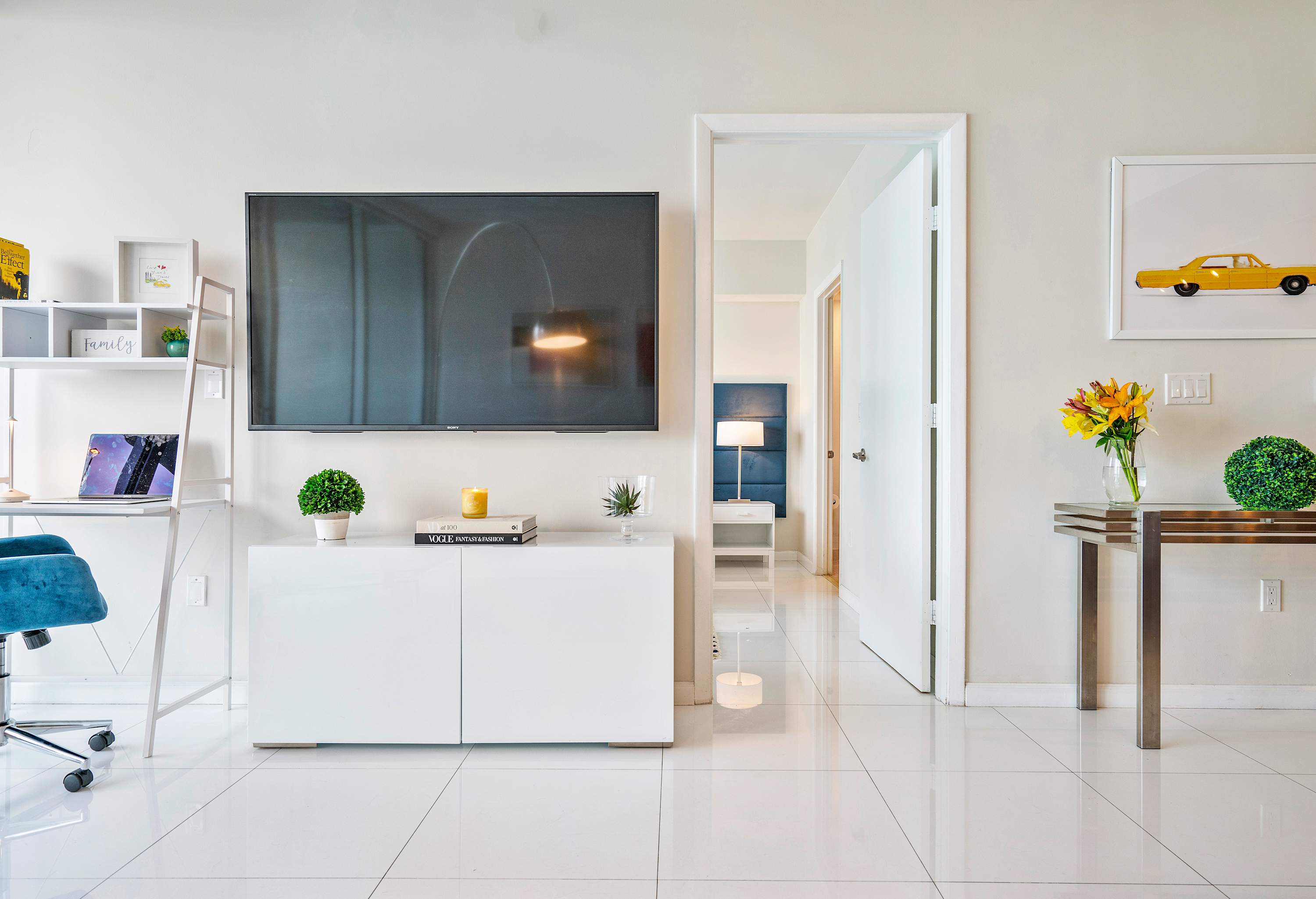 Bright living room wall featuring a wall-mounted flat-screen TV above a white console, a white shelving unit with a desk, and an open doorway leading to the bedroom.