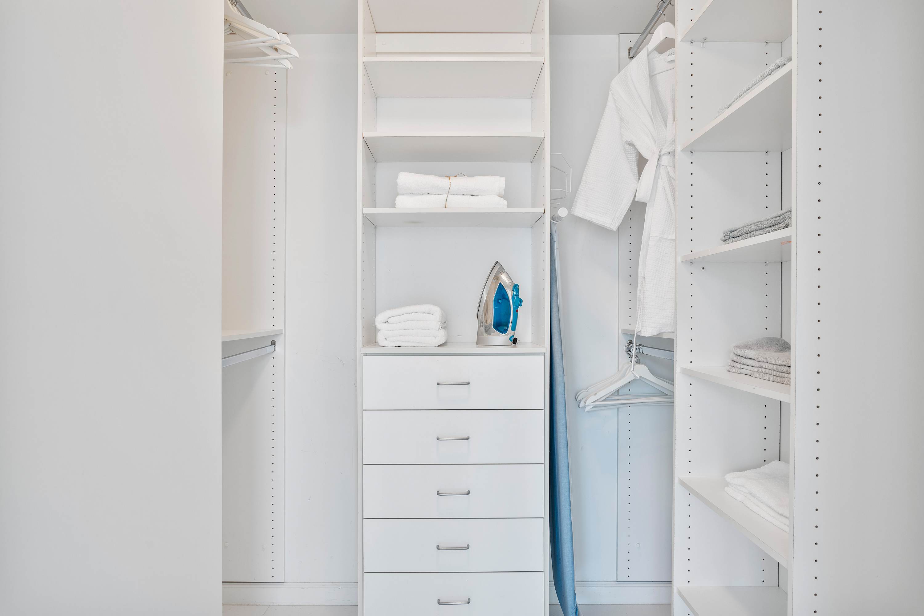 Organized walk-in closet with white built-in shelving units, multiple drawers, and hanging space, containing folded towels, an iron, and bathrobes.