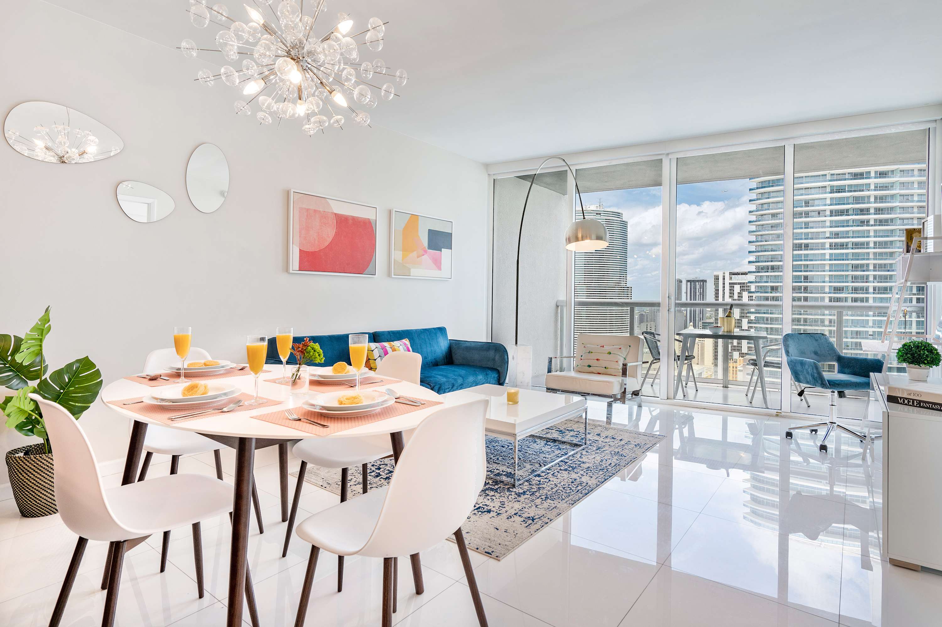 Stylish open-plan dining and living area with a round white dining table set for breakfast, a blue velvet sofa, and large windows overlooking the city skyline.