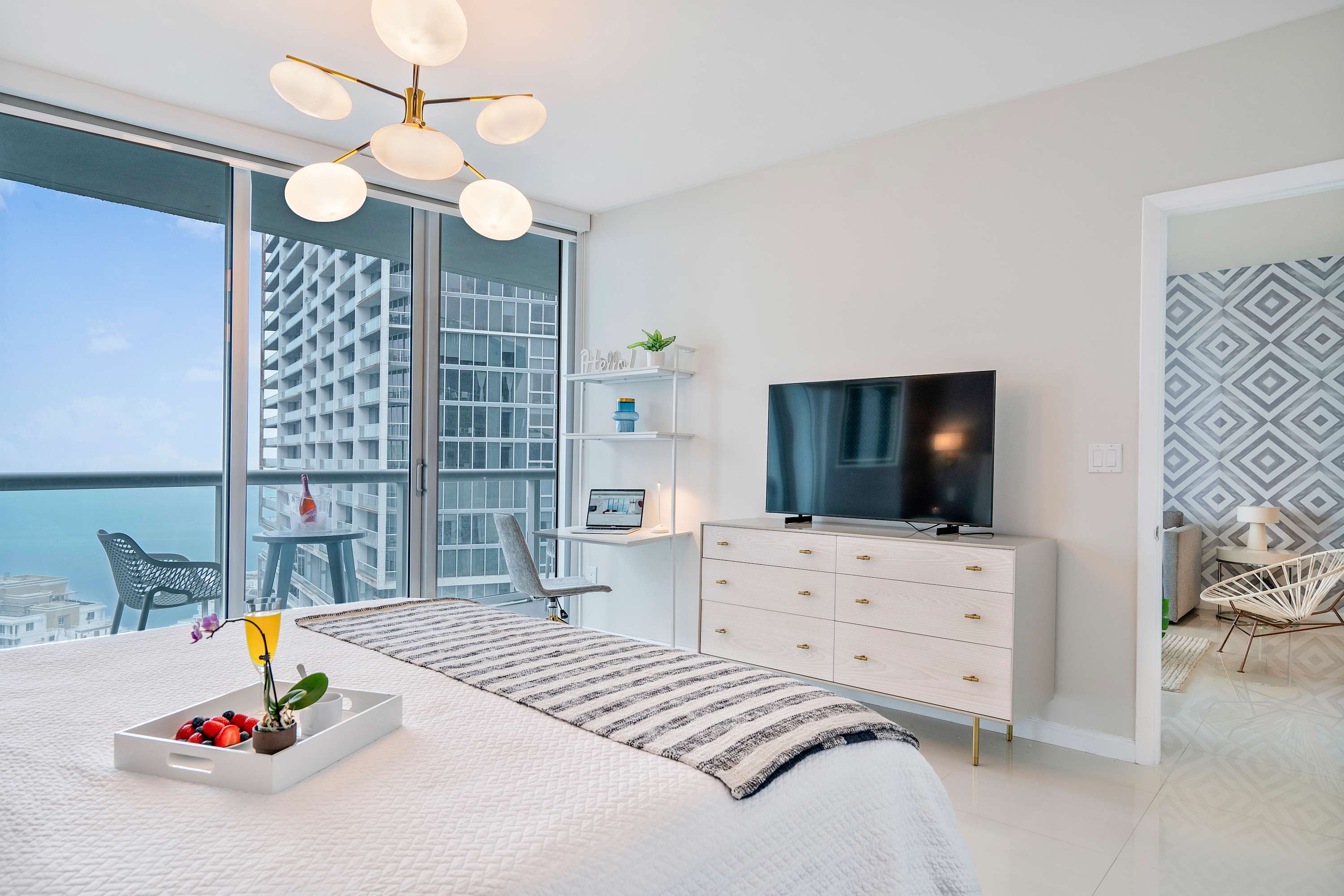 Modern bedroom with a white bed, striped throw, and a light wood dresser with a TV. A gold chandelier hangs above a small desk area, and floor-to-ceiling windows offer city and water views from a private balcony.