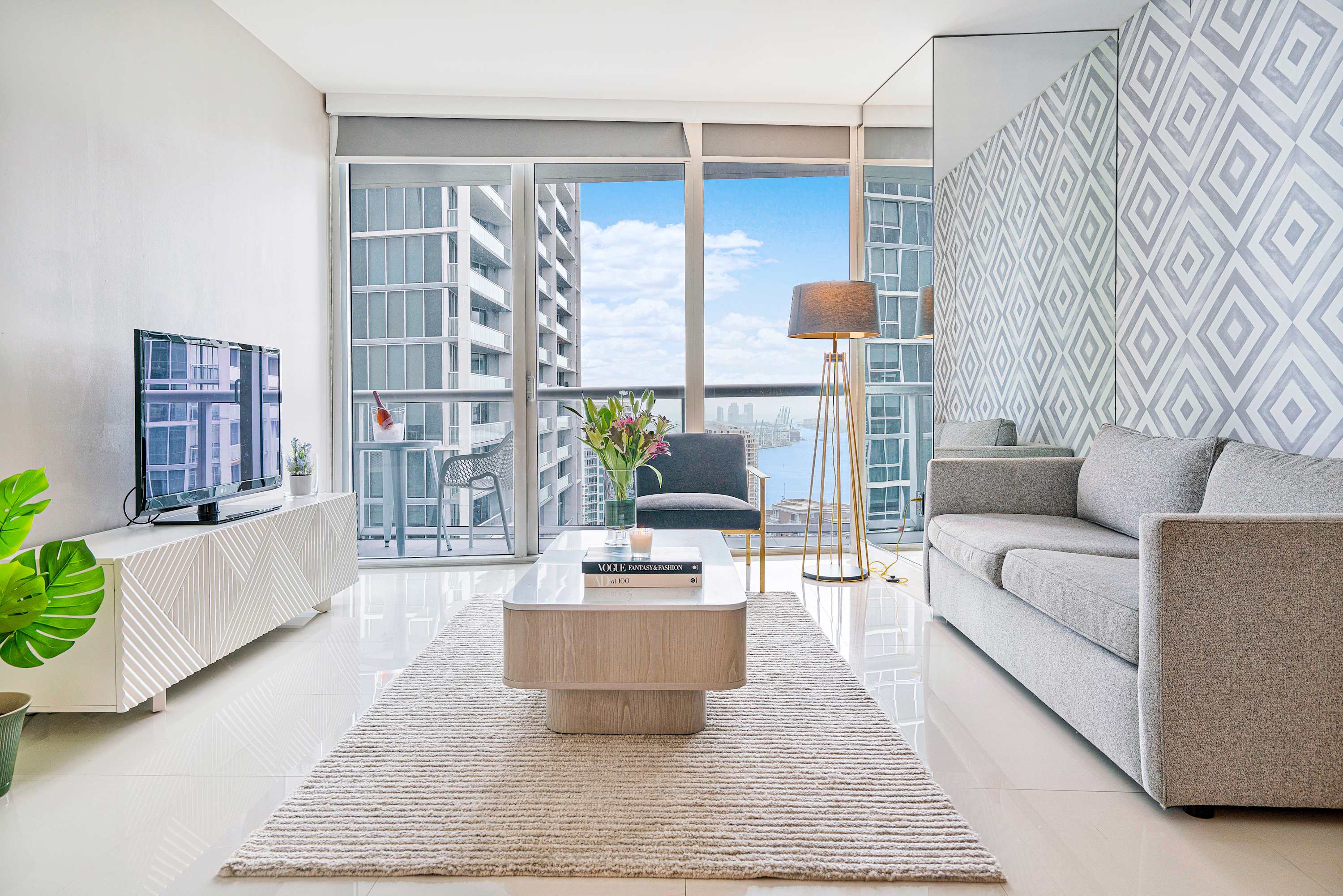 Contemporary living room featuring white tiled floors, a gray sofa, a light wood coffee table, and a patterned accent wall, with a TV console and floor-to-ceiling windows offering city and water views.