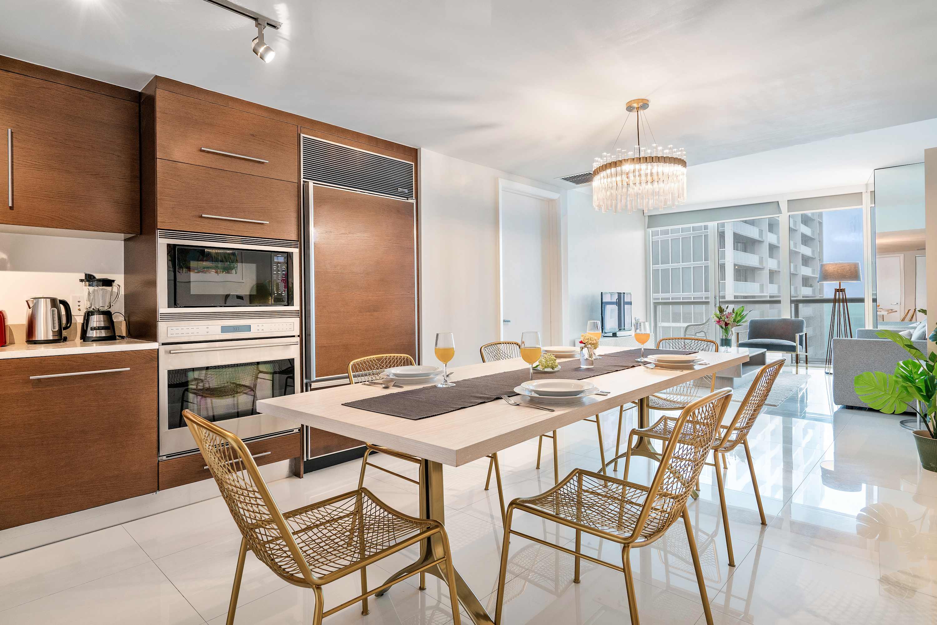 Elegant dining area with a long light wood table and gold mesh chairs, illuminated by a modern chandelier, adjacent to a sleek kitchen with wood cabinetry and integrated appliances.