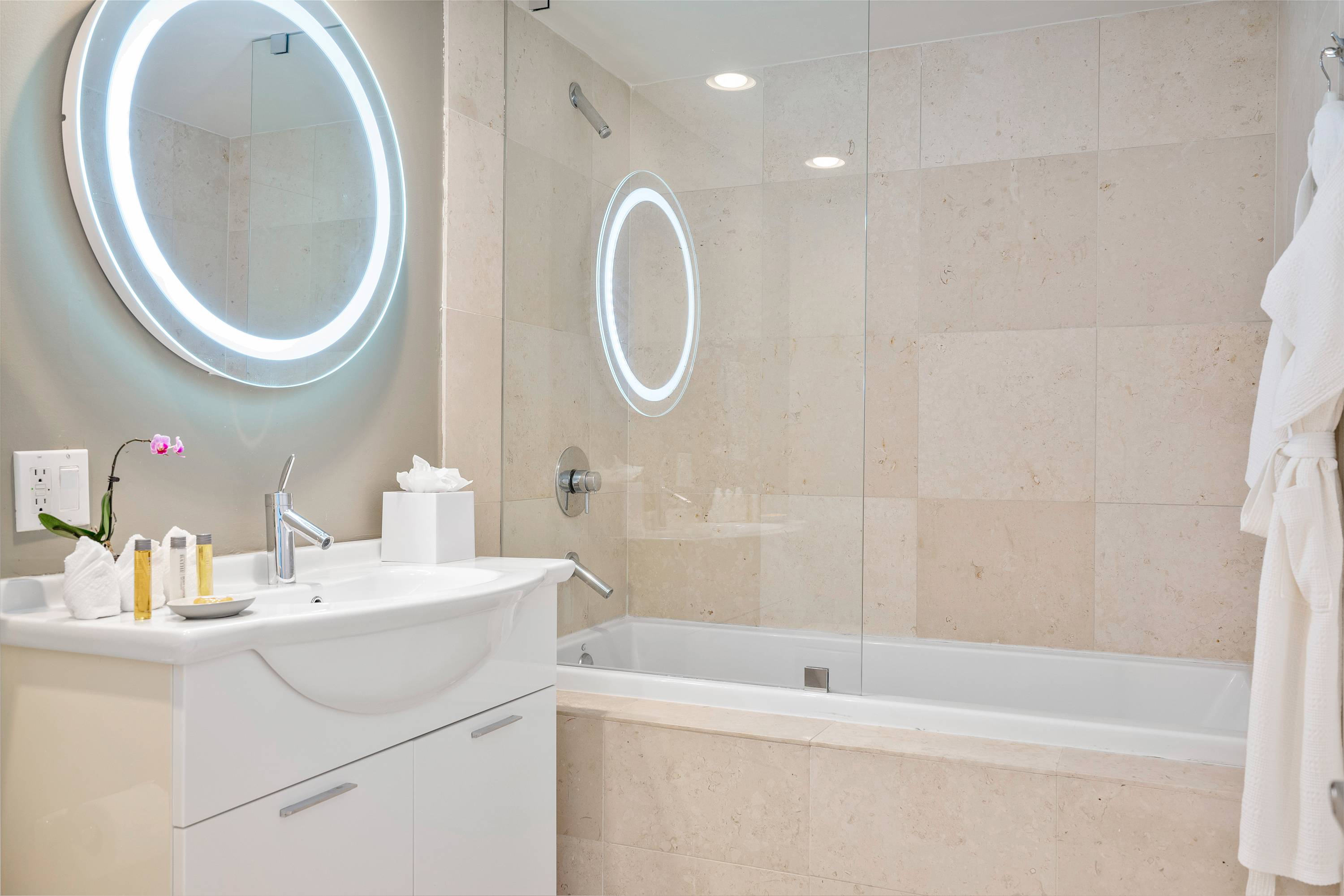 Bright bathroom featuring a white vanity with a modern sink and a large circular LED-lit mirror. A glass enclosure separates the shower/bathtub area, which is tiled with light beige stone.