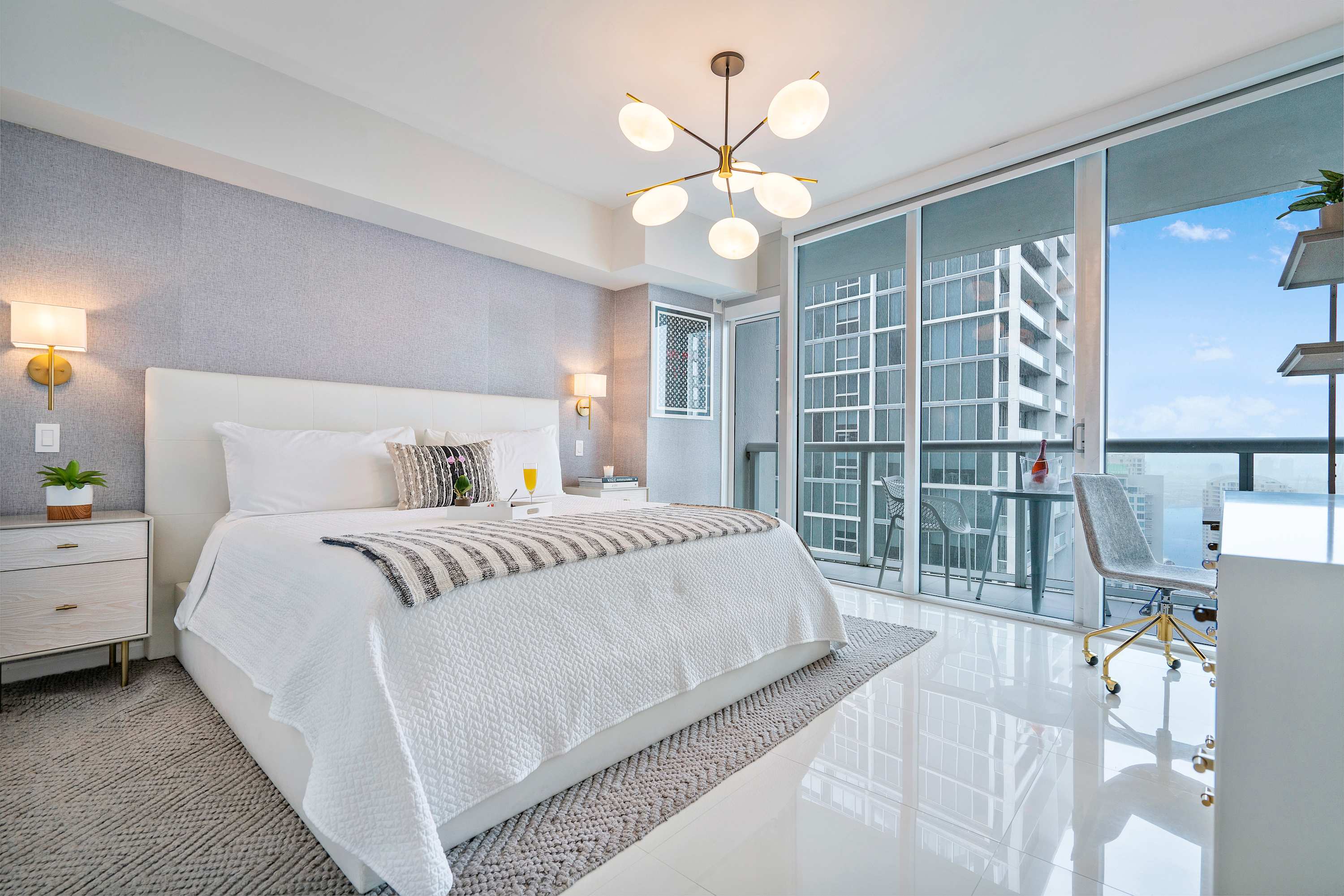 Bright bedroom with a white king-sized bed, light gray textured wallpaper, and a modern chandelier, featuring a sliding glass door leading to a private balcony with city views.