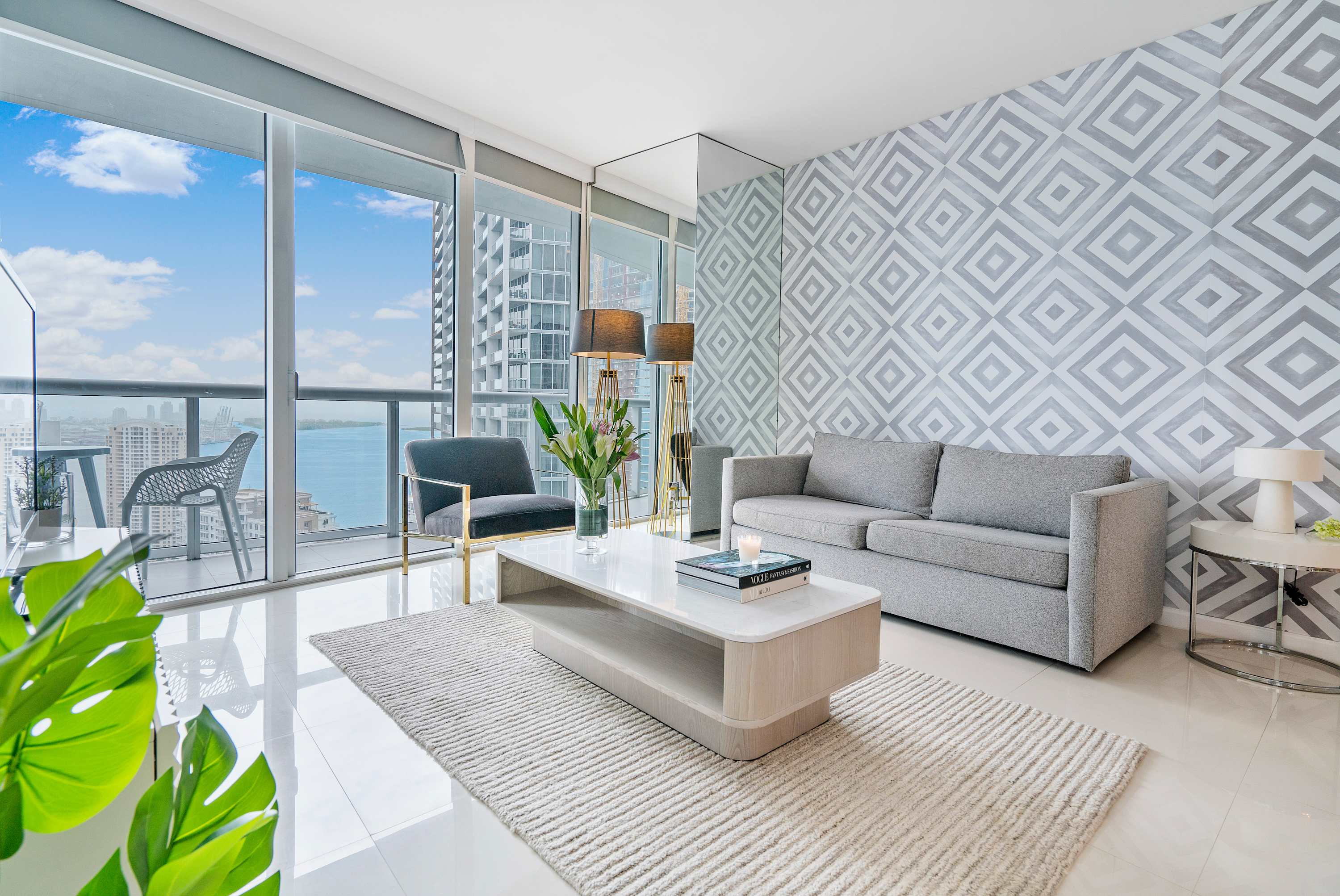 Modern living room with white tiled floors, a gray sofa, a light wood coffee table, and a patterned accent wall, featuring floor-to-ceiling windows leading to a balcony with city and water views.