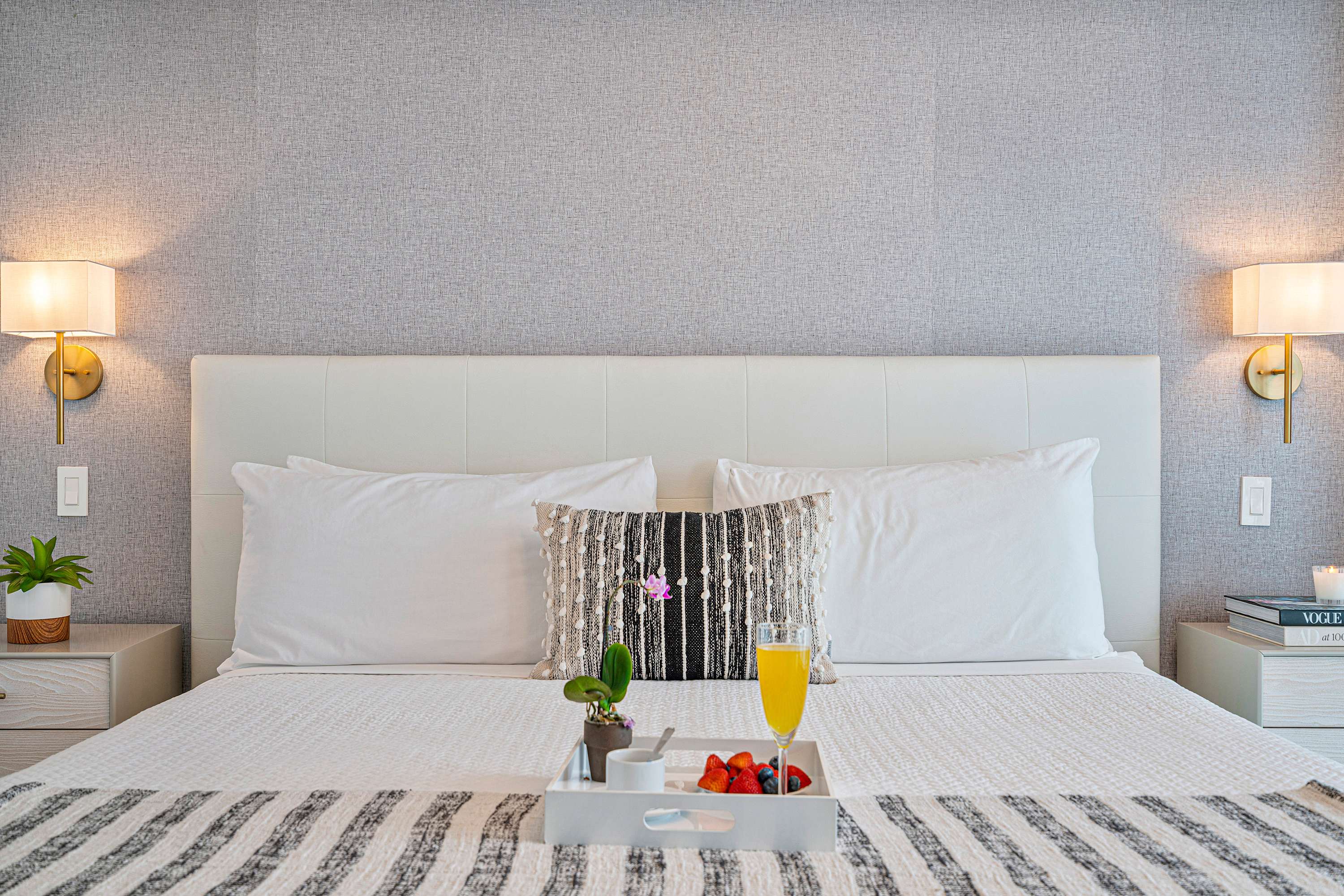 Close-up of a king-sized bed with a white upholstered headboard, white bedding, and a decorative black and white throw pillow. Two gold wall sconces flank the bed, with light wood nightstands holding a plant and books.