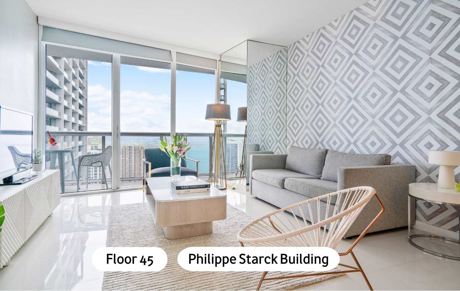 Modern living room with white tiled floors, a gray sofa, a light wood coffee table, and a patterned accent wall, featuring floor-to-ceiling windows leading to a balcony with city and water views.