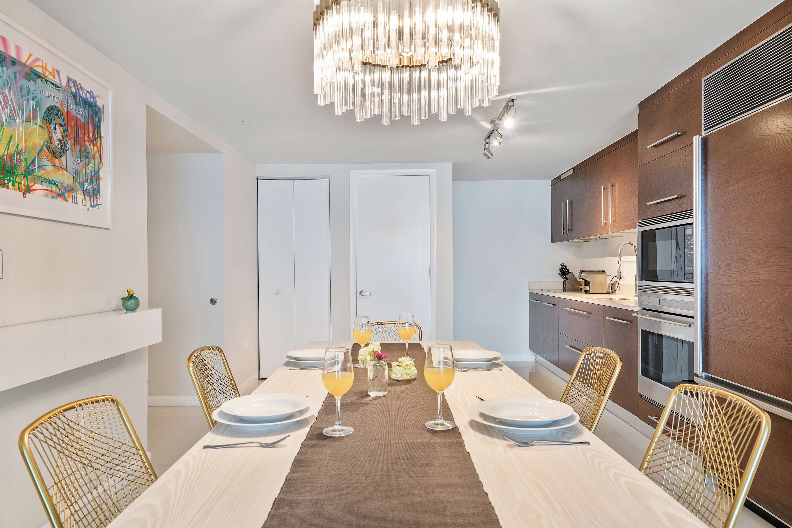 Stylish dining area featuring a long light wood table set with place settings and gold mesh chairs, under a modern crystal chandelier, with a view into the adjacent kitchen.