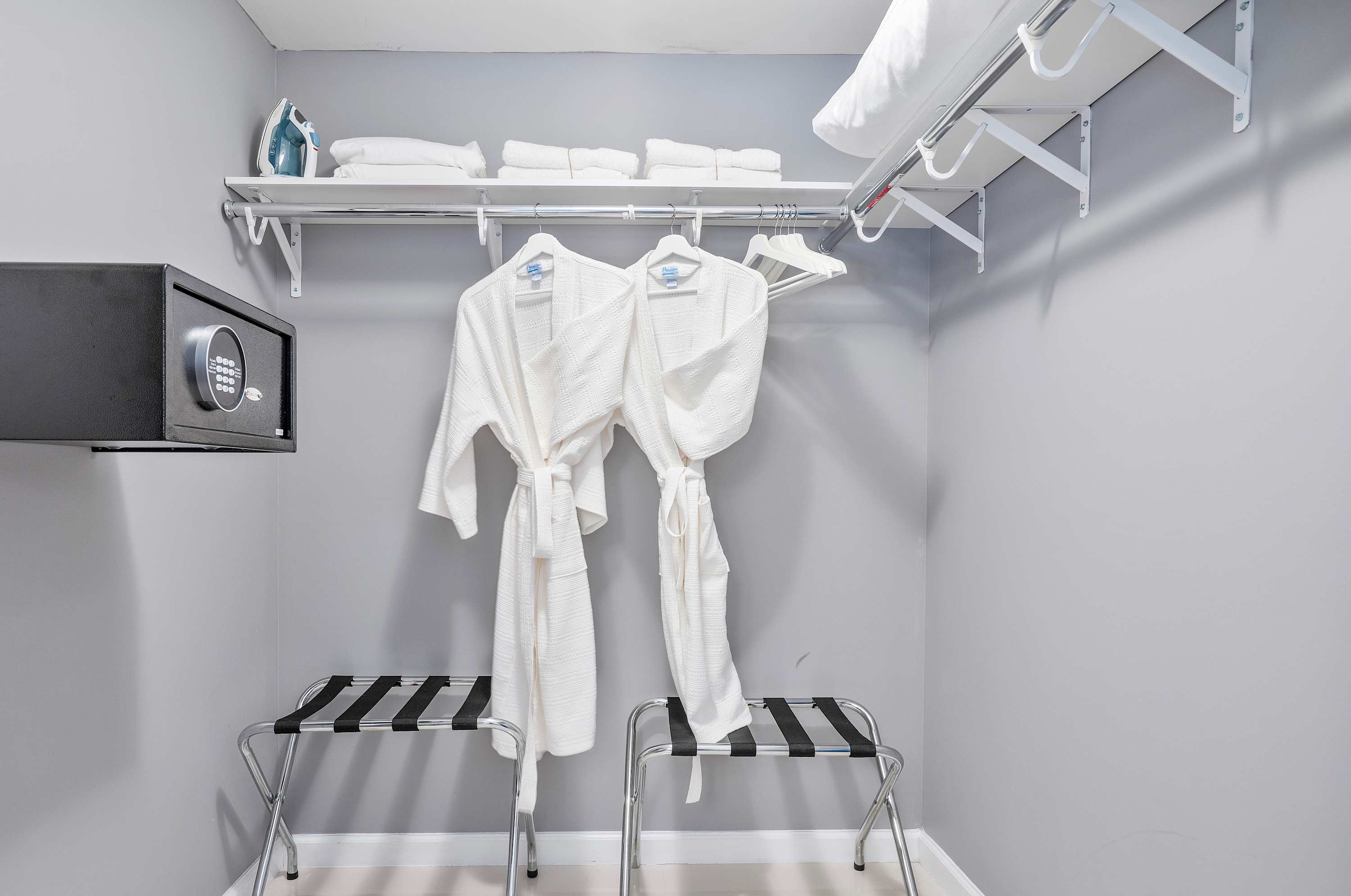 Organized closet space with two white bathrobes hanging, two luggage racks, and a wall-mounted safe. Shelves above hold folded white towels and an iron.