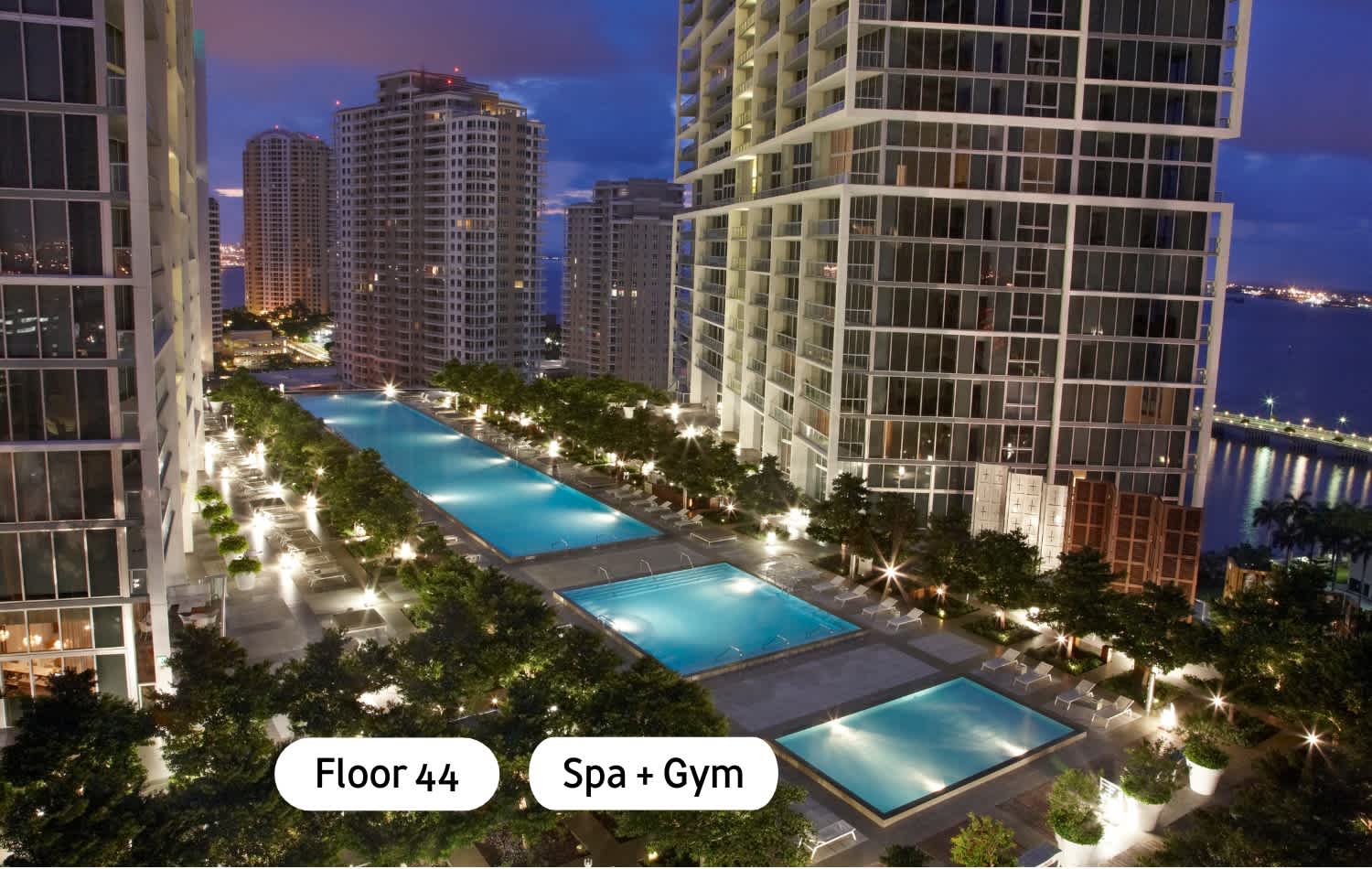 An evening aerial view showcases a long, illuminated outdoor swimming pool area with multiple pools, surrounded by lounge chairs and lush landscaping. Tall, modern high-rise buildings with glowing windows frame the scene against a twilight sky.