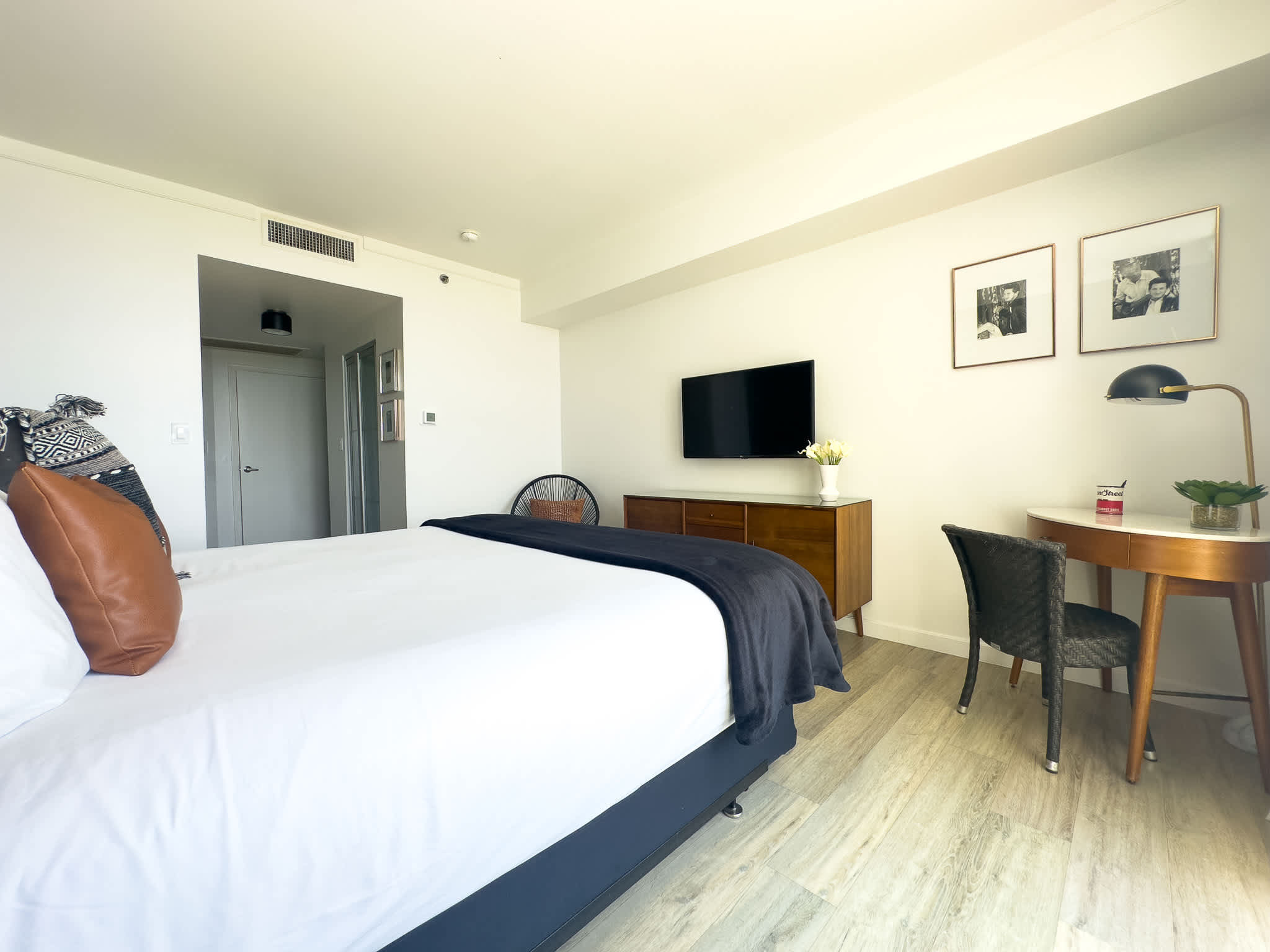A spacious bedroom features a neatly made king bed, a wall-mounted flat-screen TV above a wooden console, and a dedicated workspace with a round desk and chair, all illuminated by natural light.