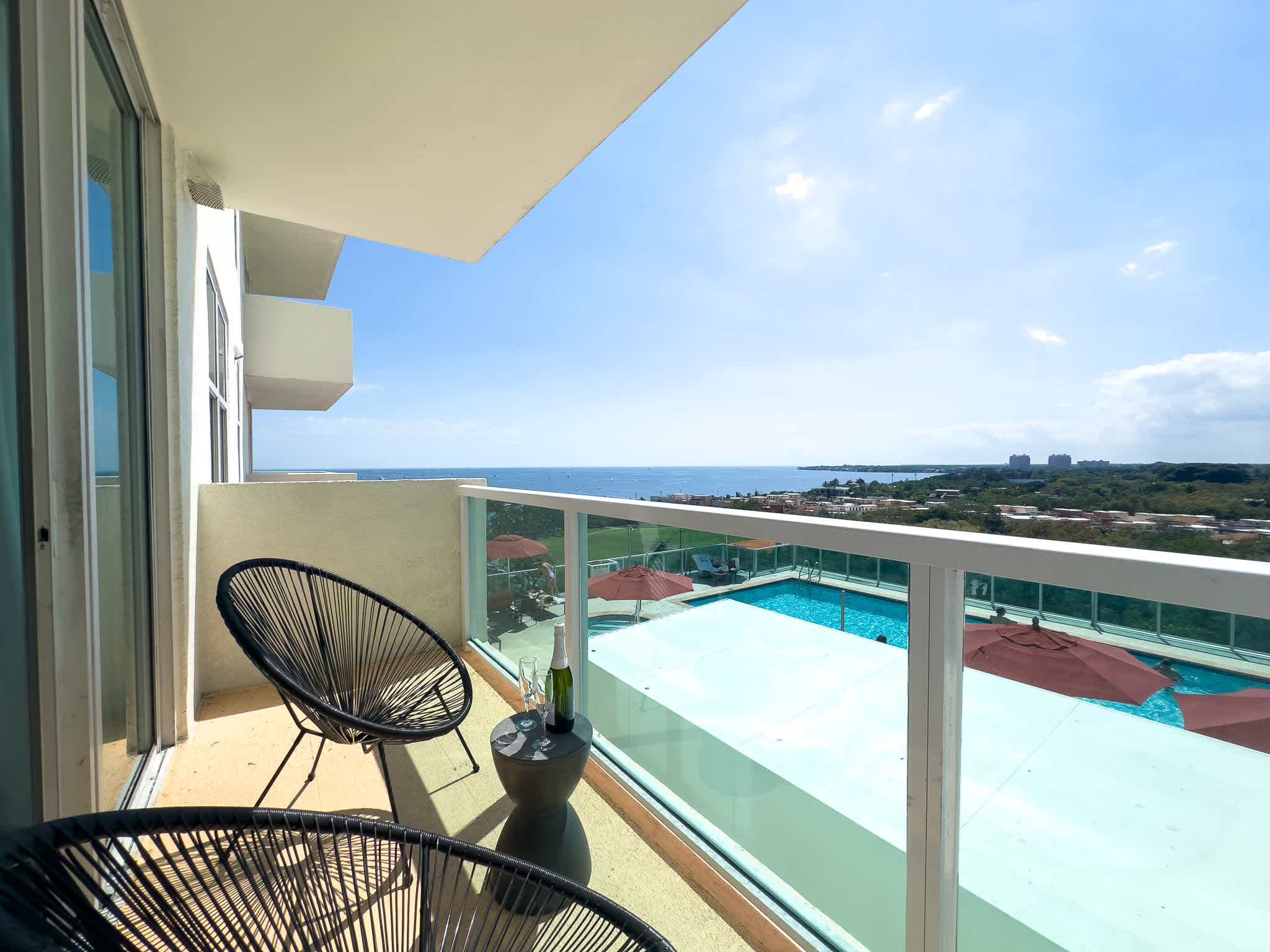 A modern balcony features two black woven chairs and a small table with a bottle and glasses, overlooking a vibrant blue swimming pool and the expansive ocean under a clear sky.