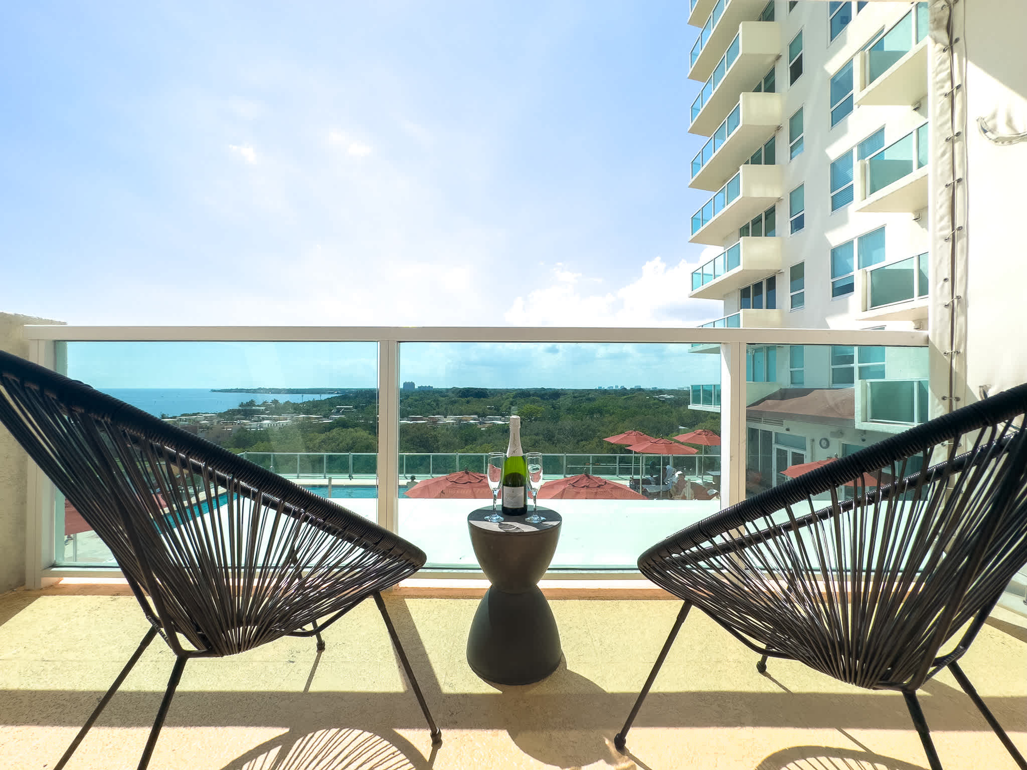 A bright balcony features two modern black woven chairs and a small table with champagne, offering a clear view of the sparkling ocean, a vibrant pool area with red umbrellas, and the adjacent high-rise building.