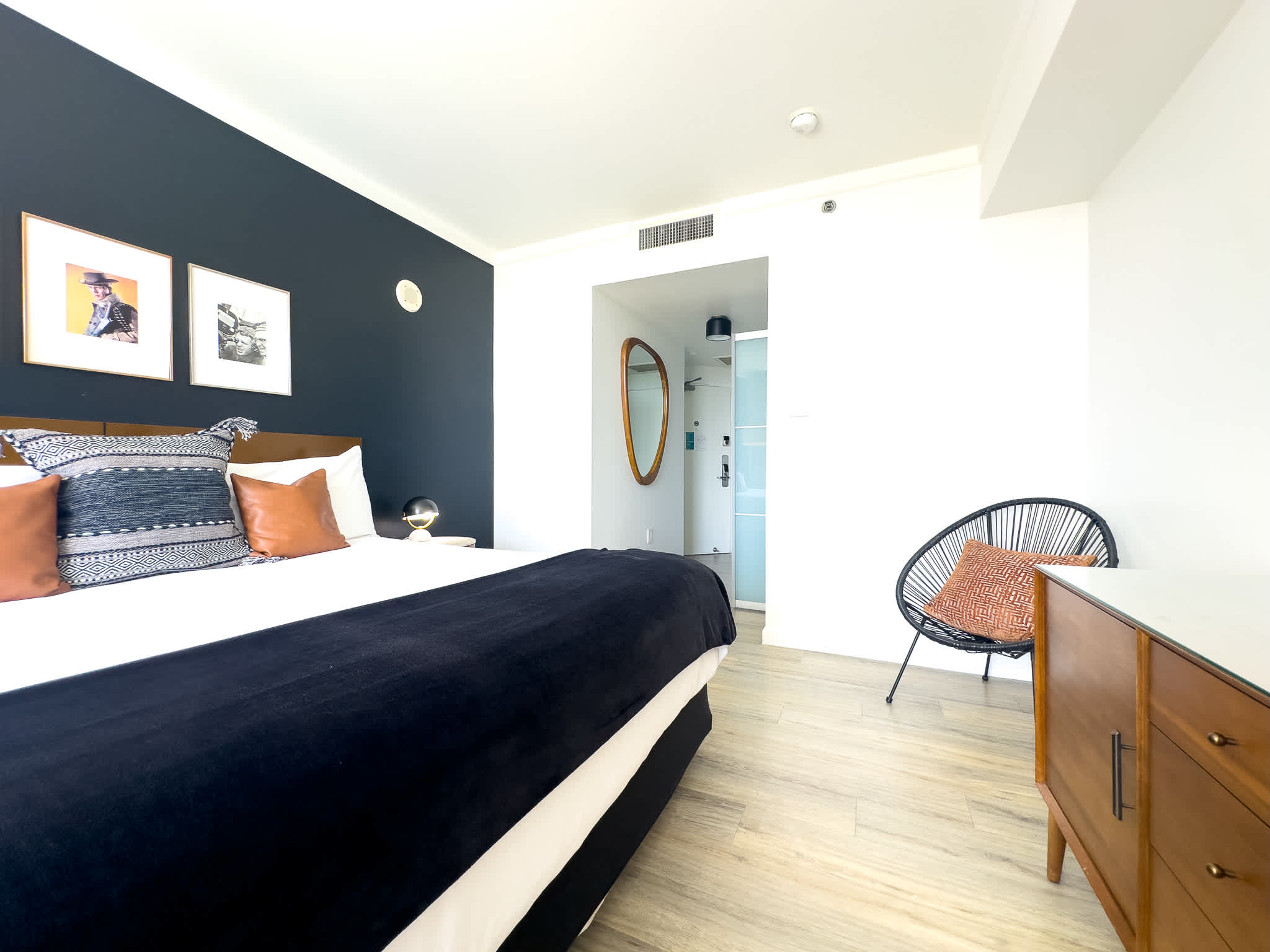 This bright bedroom showcases a king bed with a dark accent wall, a wooden dresser, a unique black woven chair, and an open doorway leading into the bathroom, all set on light wood-look flooring.