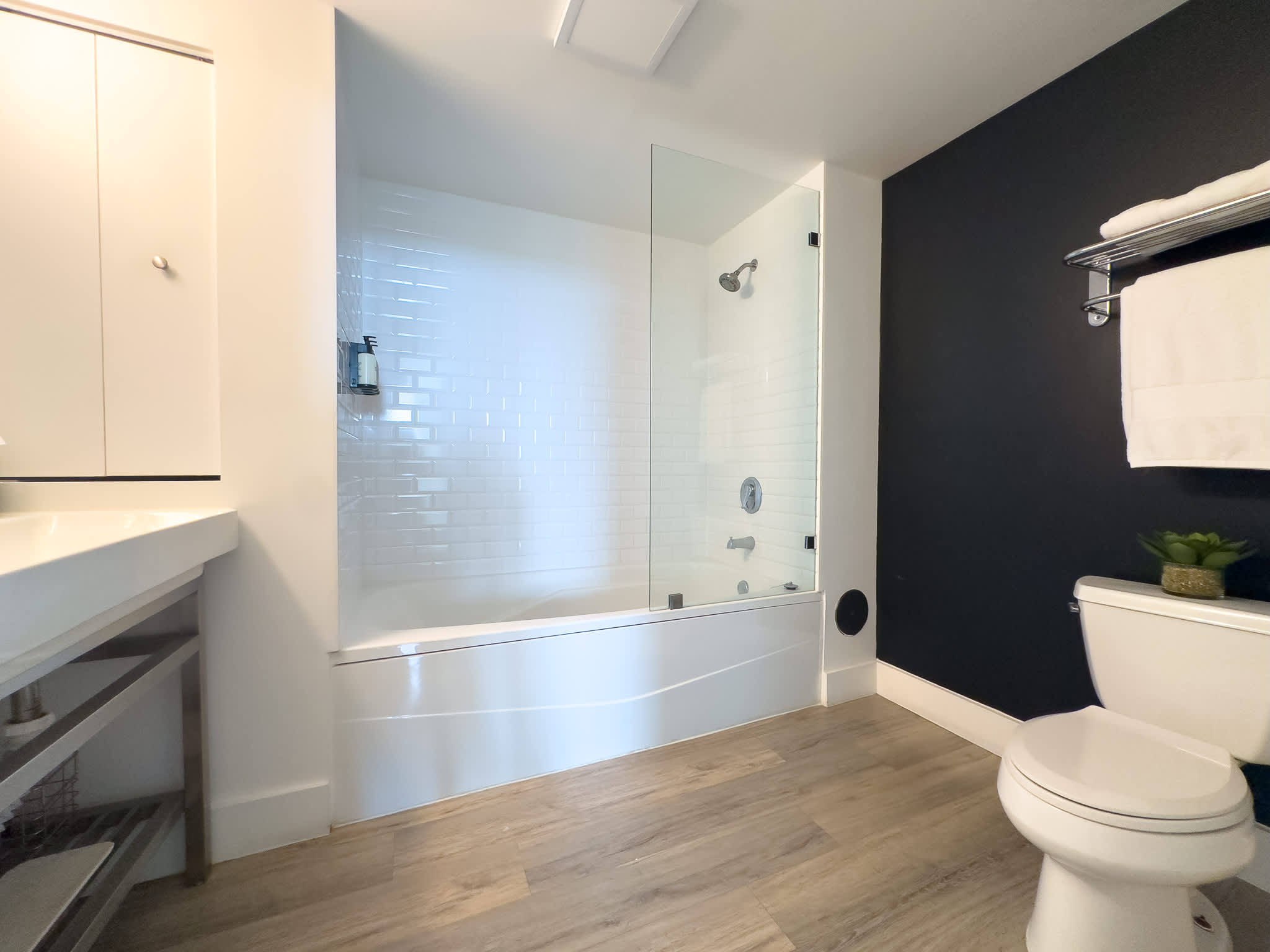 This clean bathroom includes a white bathtub with a glass shower screen, a toilet, and a dark accent wall, complemented by light wood-look flooring and bright white subway tile in the shower.