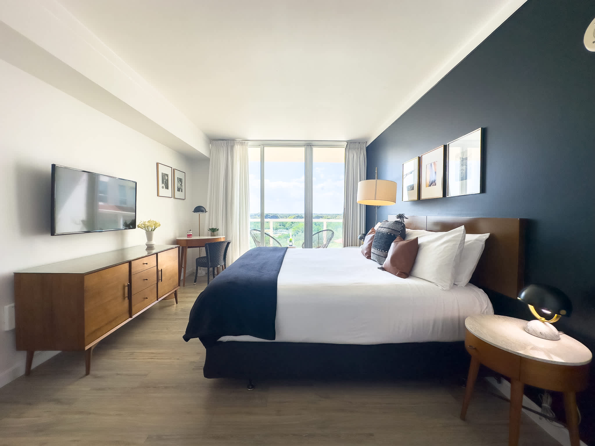 A stylish bedroom features a king-sized bed with a dark accent wall, a flat-screen TV mounted above a mid-century modern dresser, a small desk area, and floor-to-ceiling windows leading to a balcony with outdoor seating.