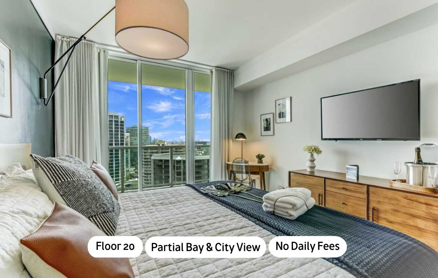 Modern bedroom featuring a large bed with white and dark blue bedding, a wall-mounted TV, and floor-to-ceiling windows leading to a balcony with city and partial bay views.