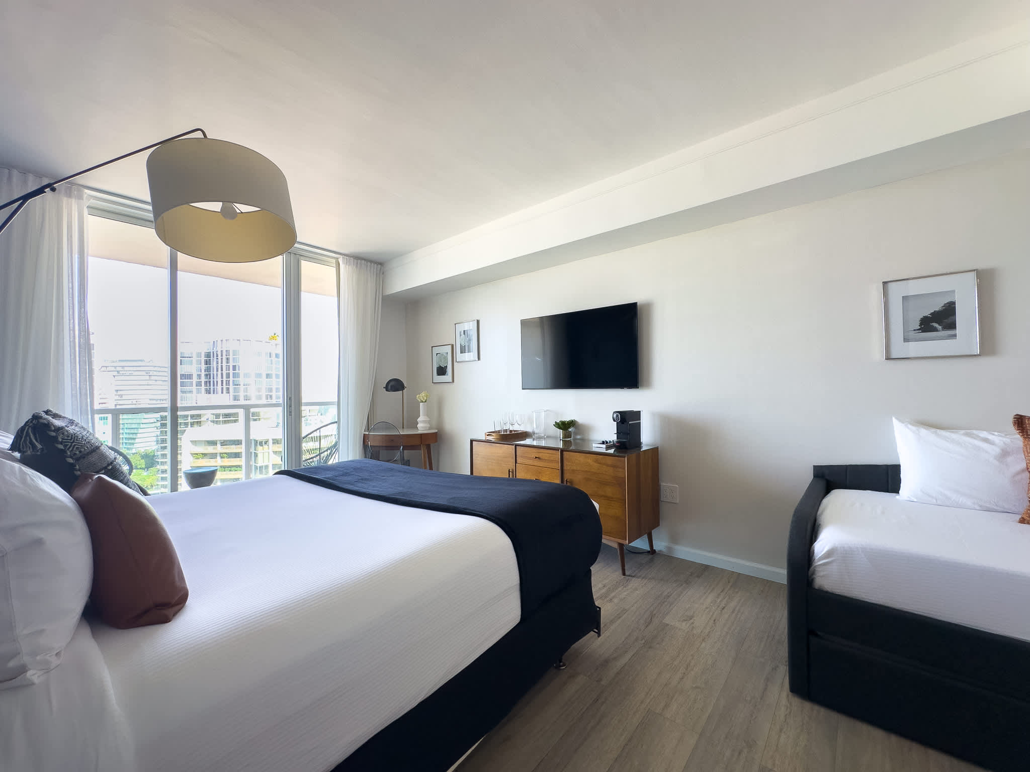 Wide view of a contemporary bedroom featuring a large bed, a daybed with an orange accent pillow, a wooden console with a TV, and a balcony with city views.