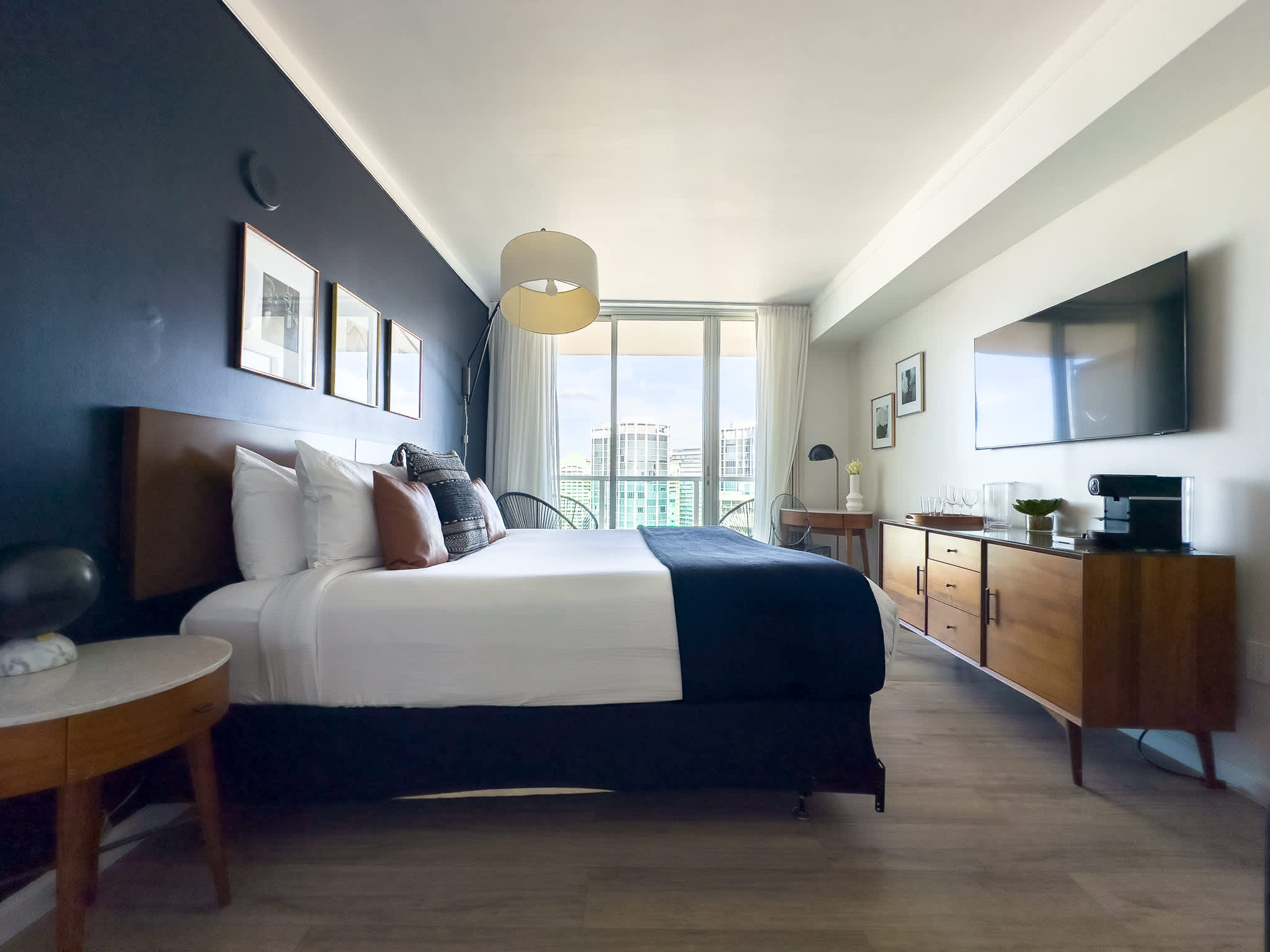 Spacious bedroom with a large bed, a long wooden dresser with a flat-screen TV mounted above it, and floor-to-ceiling windows opening to a balcony with city views.