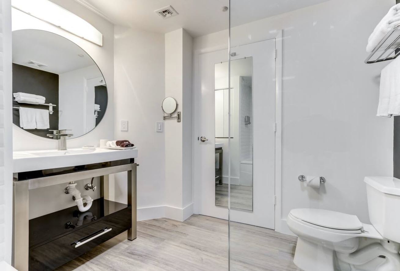 Clean and contemporary bathroom with a white toilet, a modern vanity with an open shelf design, and a large round mirror, featuring light wood-look flooring.