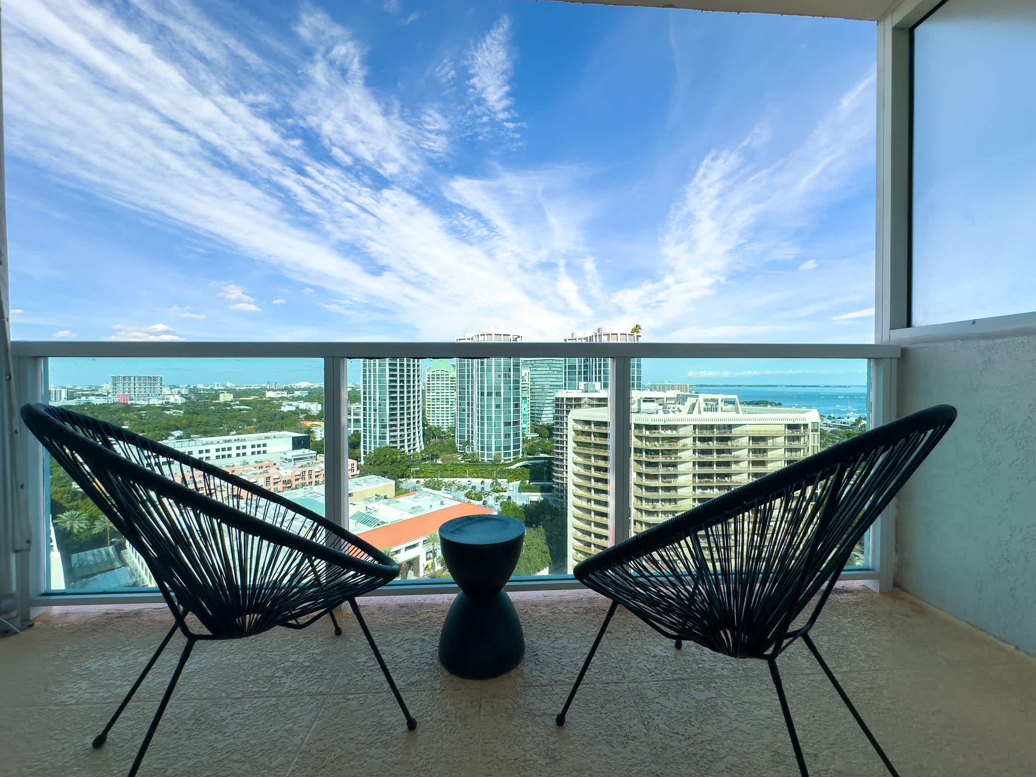 Private balcony featuring two black woven chairs and a small round table, offering expansive views of the city skyline and a distant bay under a bright blue sky.