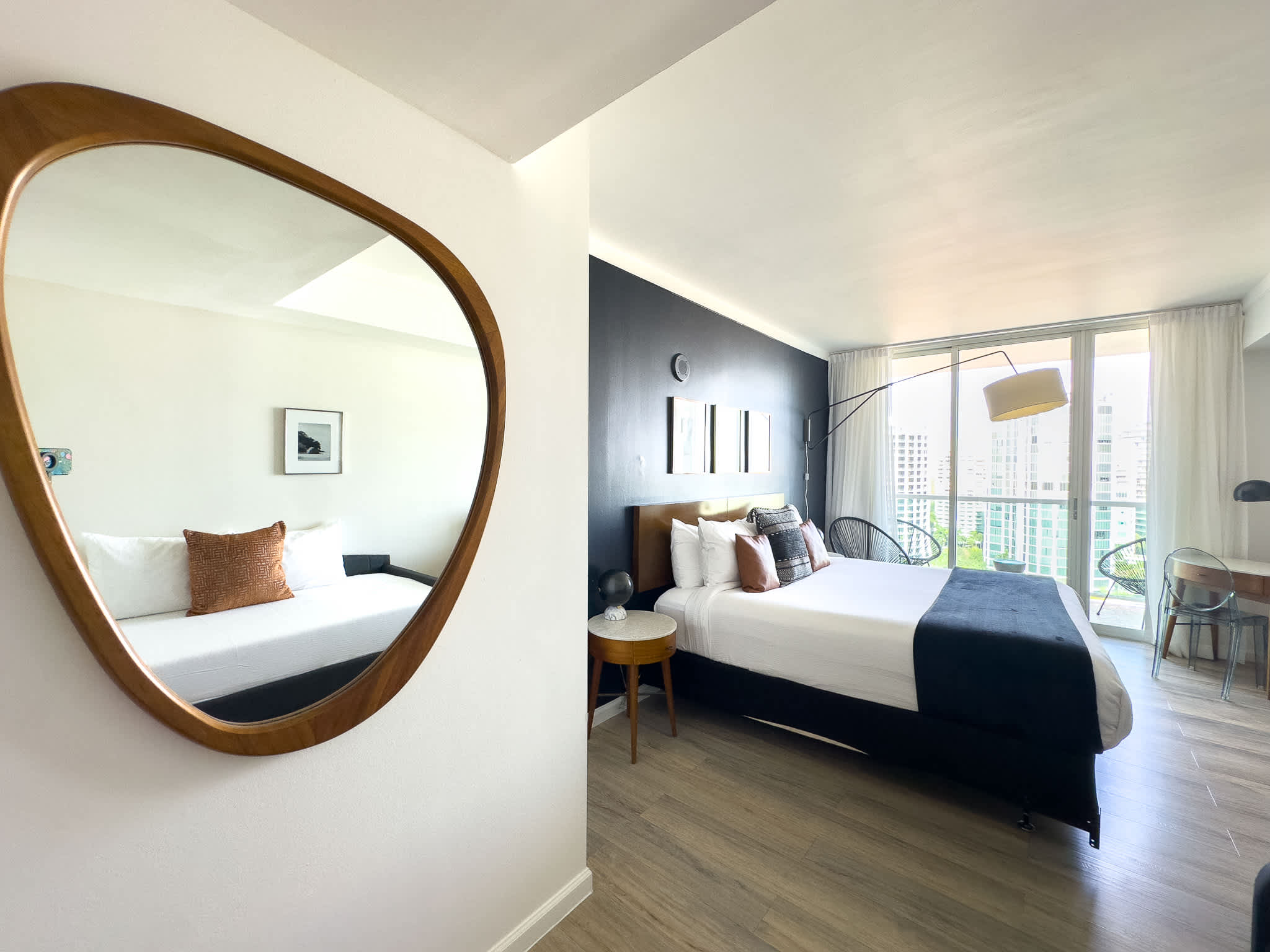 View into a modern bedroom with a large bed, dark accent wall, and a unique organic-shaped mirror reflecting the room, leading to a balcony with city views.