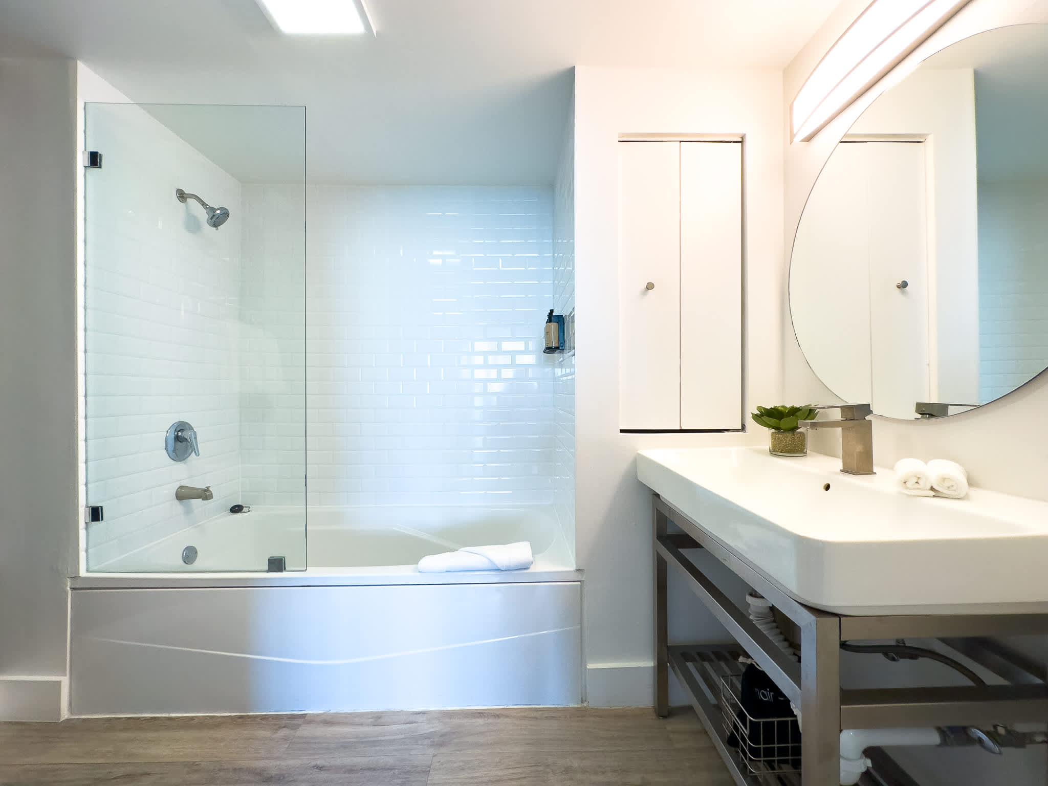 Bright and modern bathroom featuring a white subway-tiled shower/bathtub combo with a glass partition, and a sleek vanity with a rectangular sink and a large round mirror.