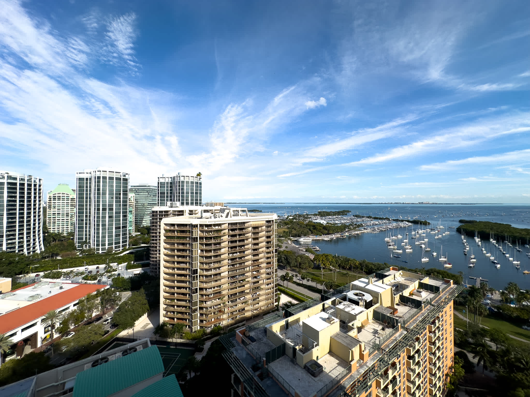 Panoramic aerial view of a vibrant city skyline with numerous high-rise buildings, overlooking a large bay filled with sailboats under a bright blue sky with scattered clouds.