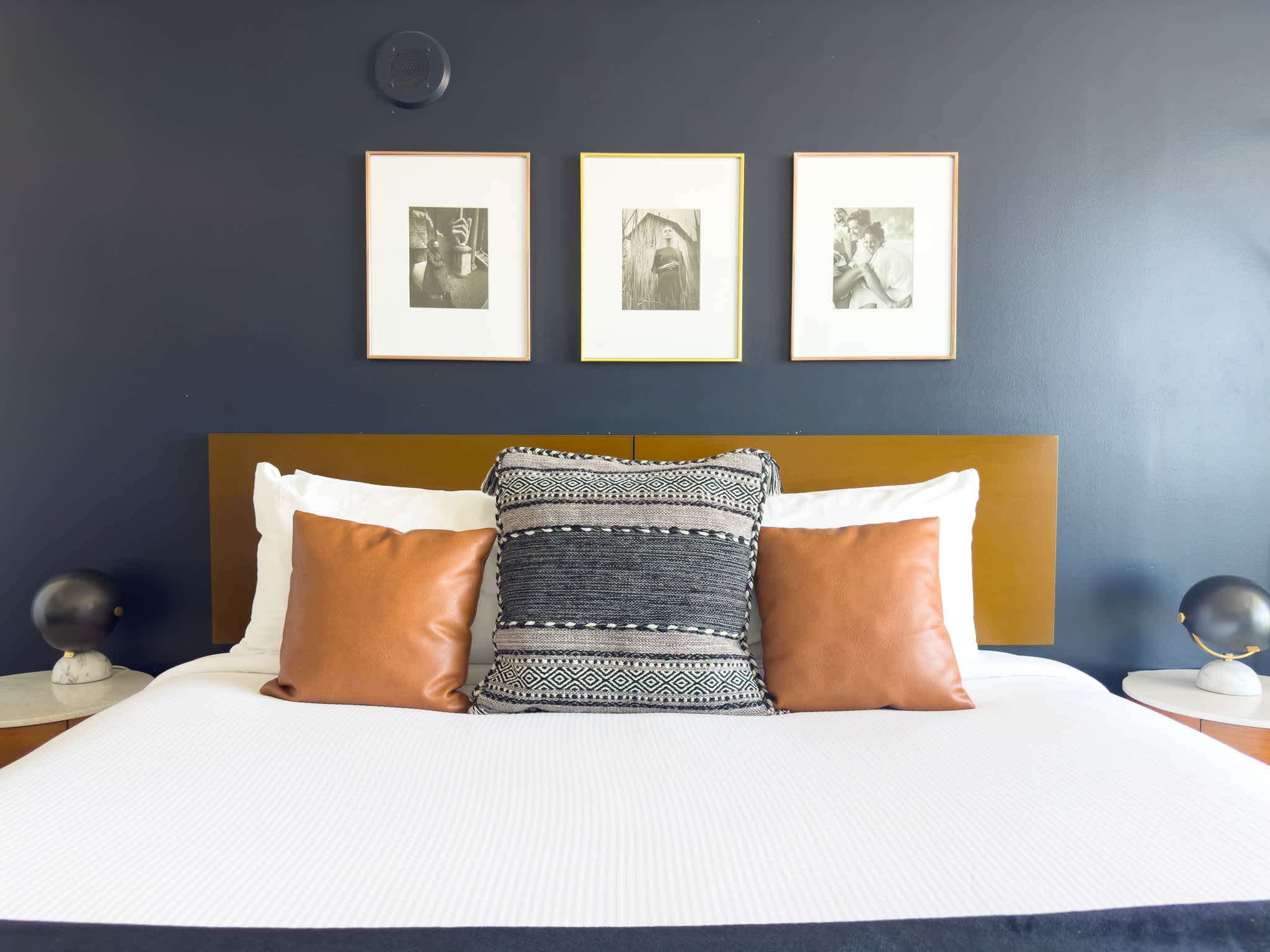 Close-up of a neatly made bed with a brown headboard against a dark blue accent wall, adorned with white pillows, a patterned throw pillow, and two brown accent pillows.