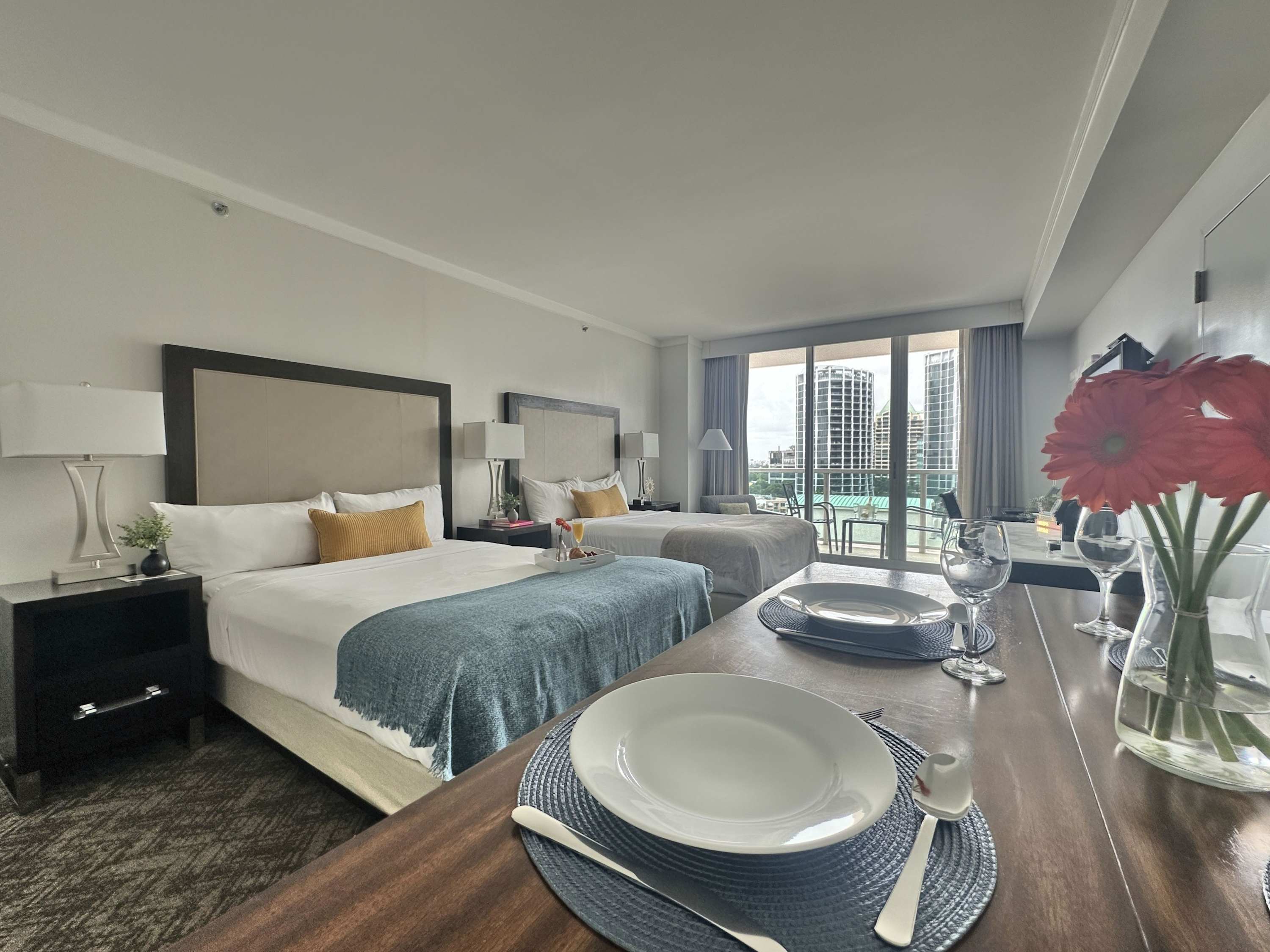 A bright studio room featuring two queen-sized beds with white linens and blue accents, a wooden dining table set for two, and a large sliding glass door leading to a balcony with a view of modern city buildings.