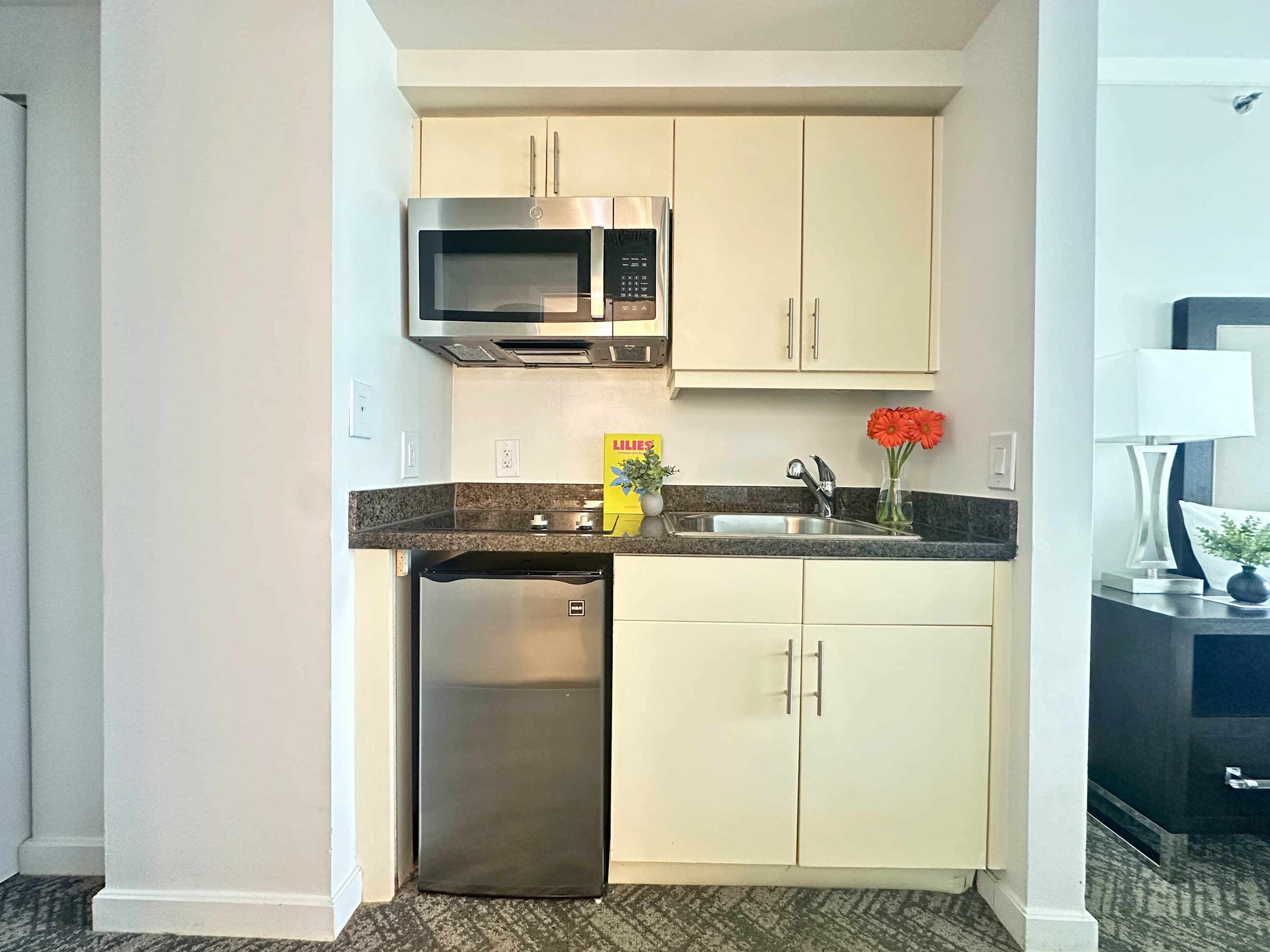 A compact kitchenette featuring light cream cabinetry, a dark granite countertop, a stainless steel microwave, a sink, and a mini-fridge, with a small vase of red flowers adding a pop of color.